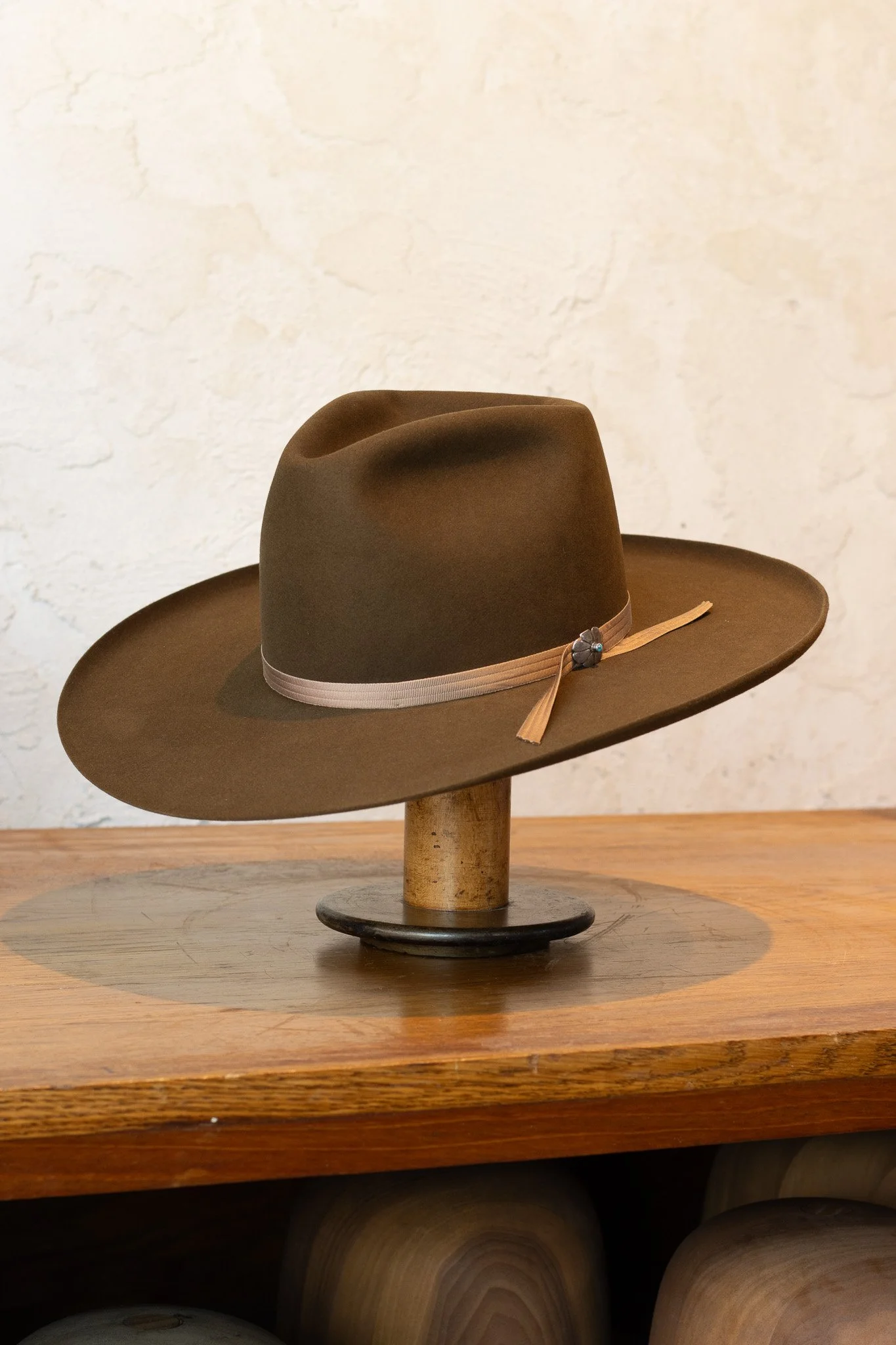Brown wide-brimmed hat with a ribbon and decorative element, displayed on a wooden stand on a rustic wooden table.