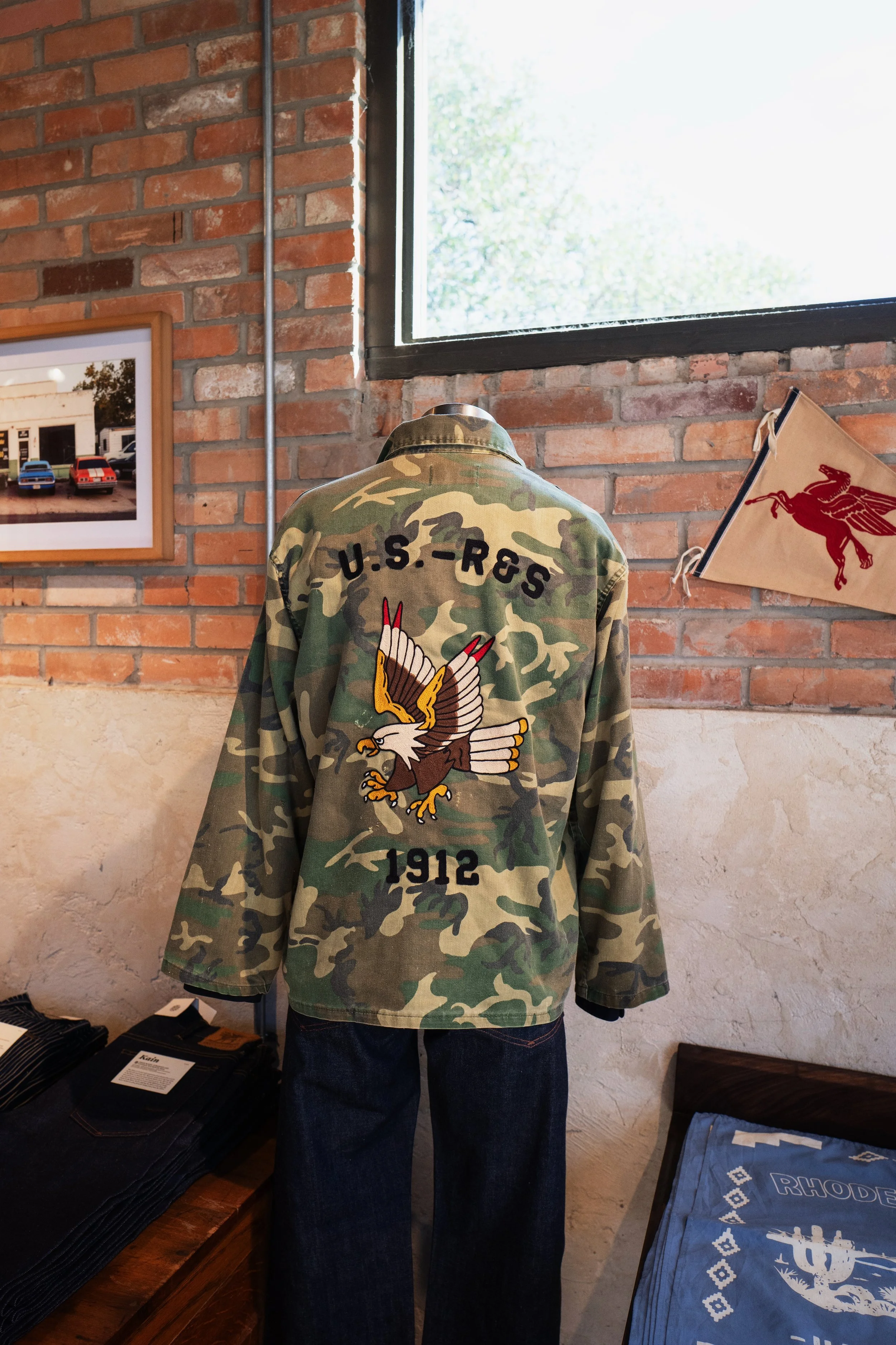 A camouflage U.S. military jacket with an eagle patch and the year 1912 displayed on a mannequin. The jacket is displayed in a room with brick walls, a window, and framed photos. A small pennant banner and a folded cloth with the Rhode Island emblem 