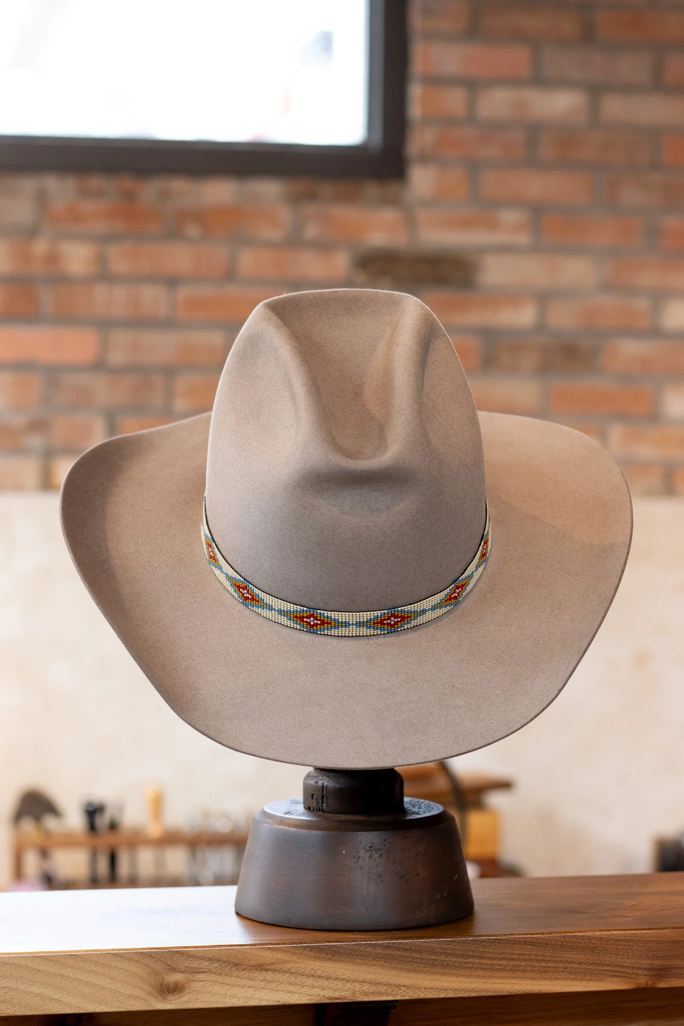 A beige cowboy hat with a colorful beaded band displayed on a black stand on a wooden surface, with a brick wall and window in the background.