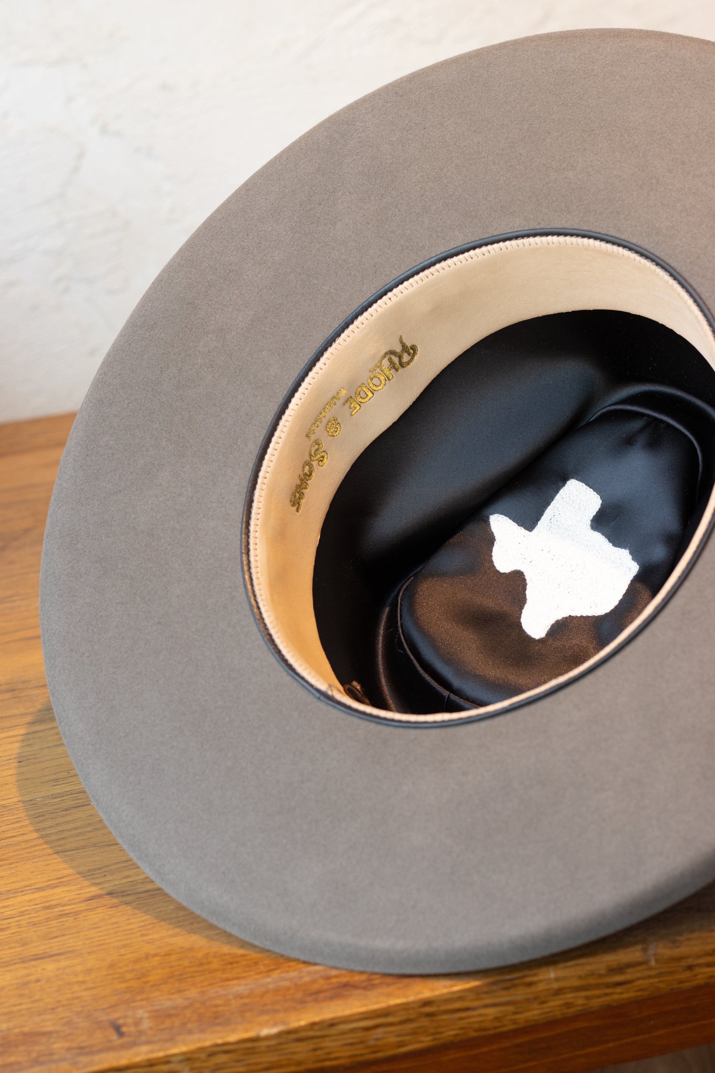 Gray fedora hat with a black interior lining and a white logo, resting on a wooden surface.