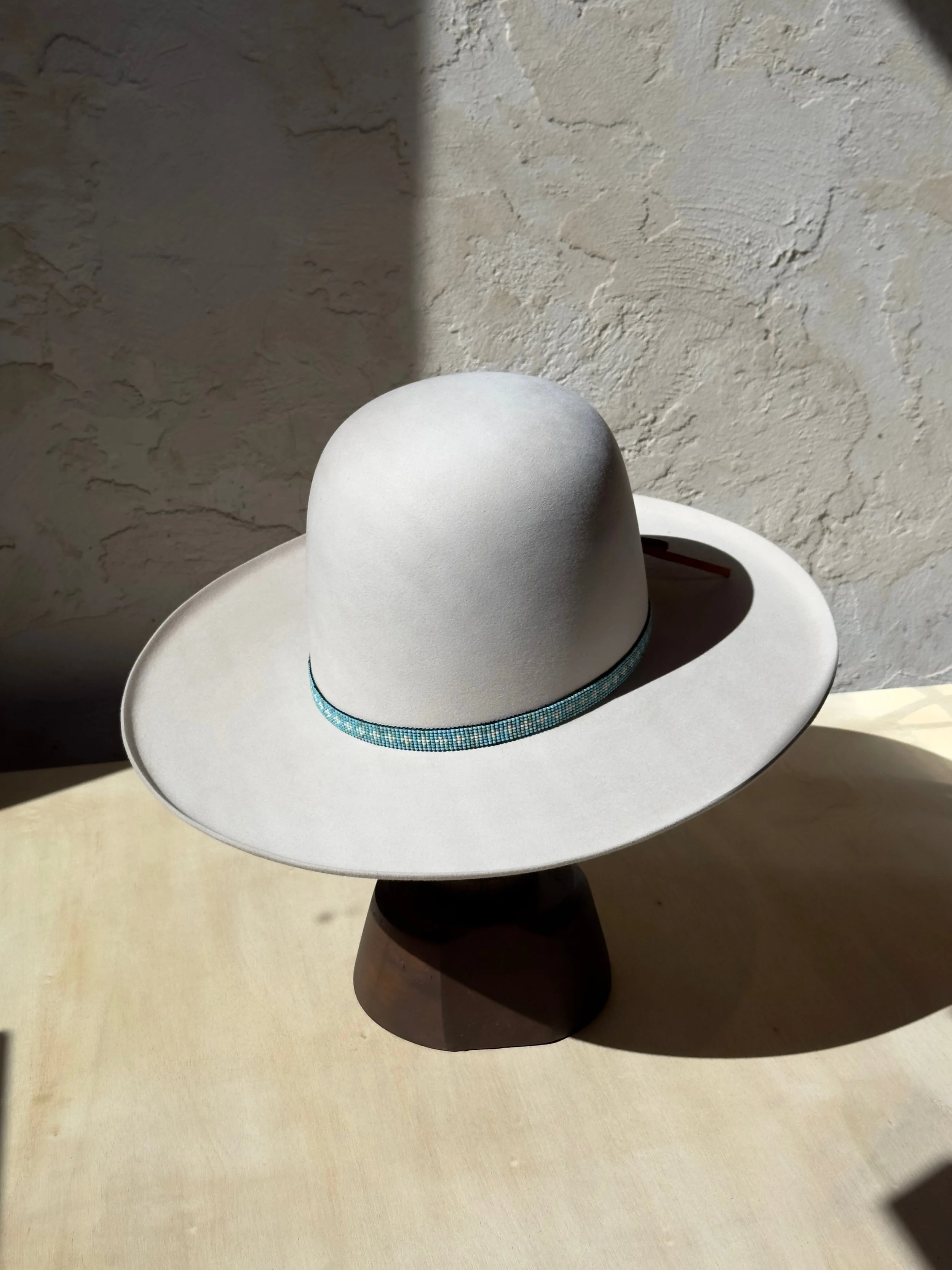 A white wide-brimmed hat with a decorative blue band, sitting on a dark stand, casting a shadow on a textured beige wall.