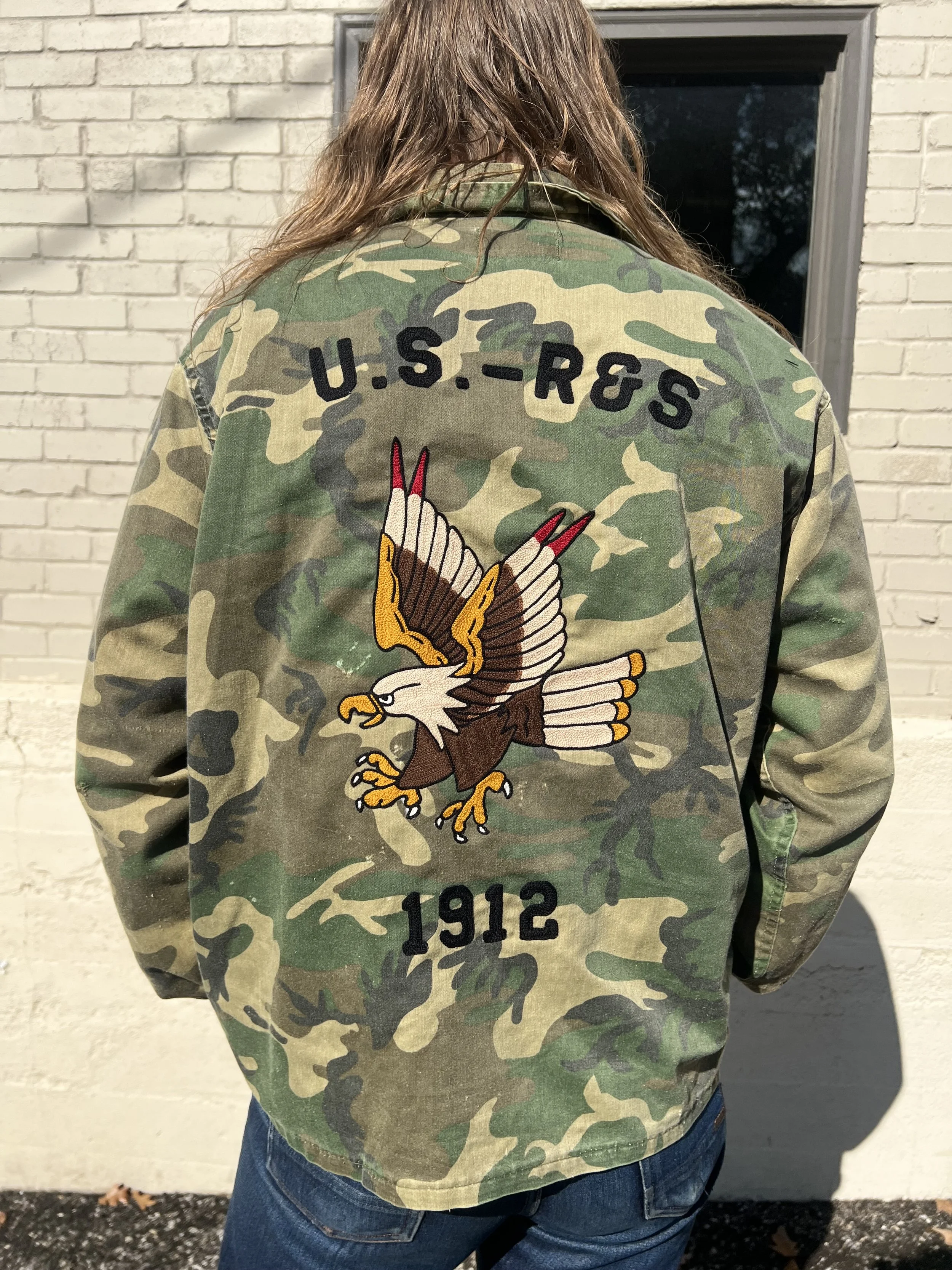 Back of a person wearing a green camo jacket with an embroidered U.S. Army patch, an eagle with outstretched wings, and the year 1912.
