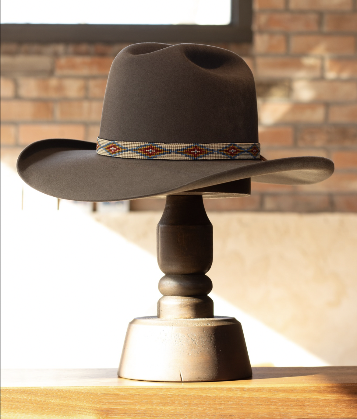 A brown cowboy hat with a beaded band resting on a wooden stand, placed on a table with a brick wall and window in the background.