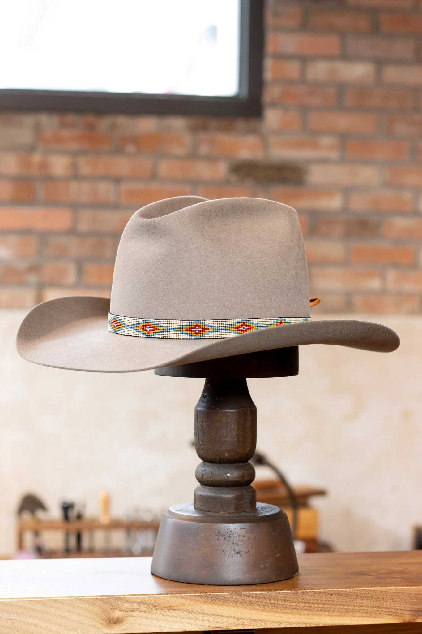 A beige cowboy hat with a colorful beaded band on a dark wooden stand on a wooden surface, with a brick wall and window in the background.