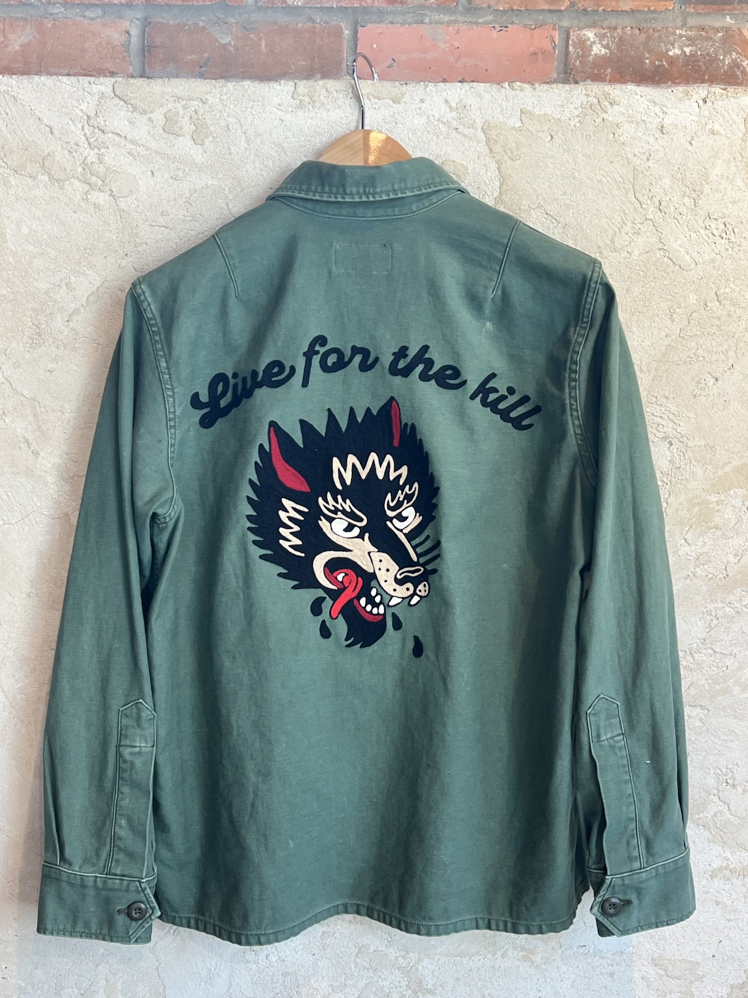 Green jacket with embroidery of a wolf's head and the words "Live for the hill" on the back hanging on a wooden hanger.