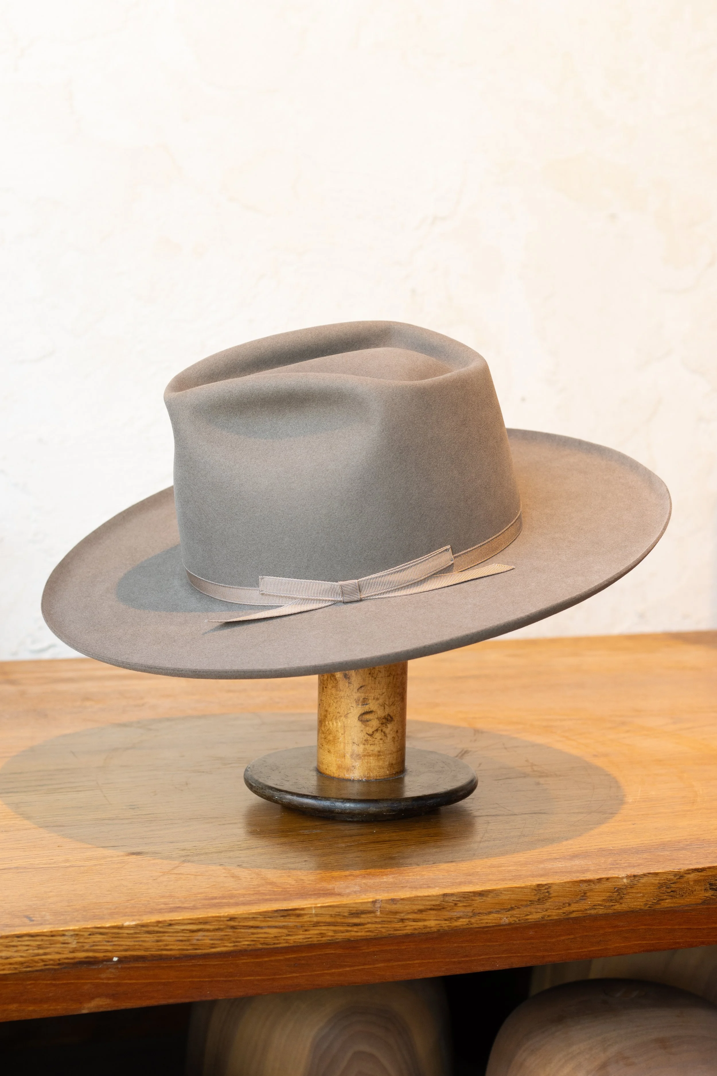 A gray fedora hat with a ribbon is placed on a wooden display stand on a wooden surface.