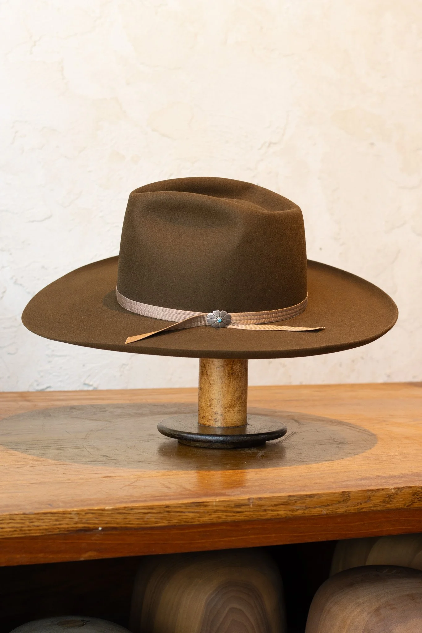 Brown wide-brimmed hat with a cream ribbon and a small flower detail, displayed on a wooden stand on a wooden table.