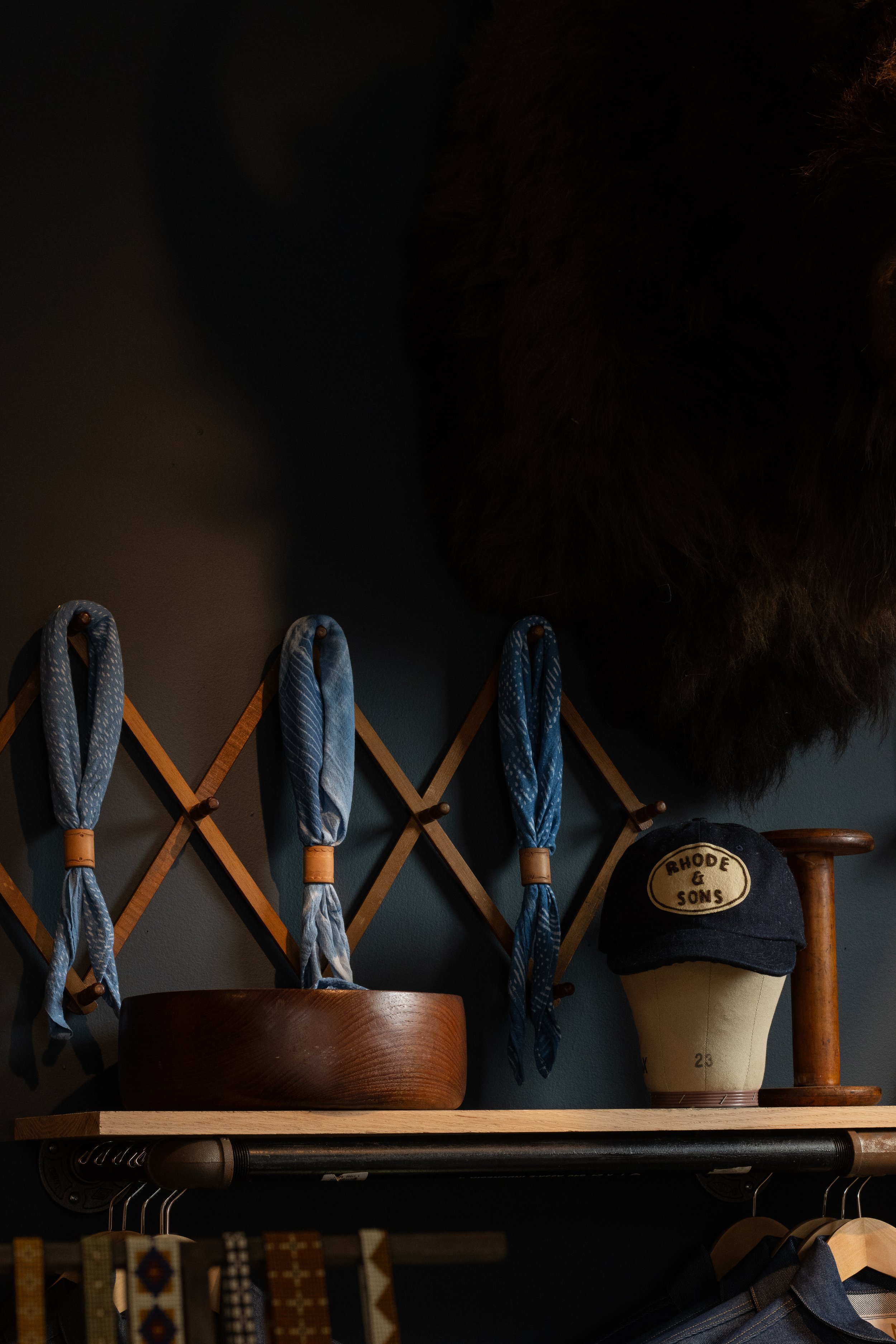 Four folded blue scarves hanging on a wooden wall rack with a rounded wooden bowl, a Rhode & Sons cap, and a wooden spool on a shelf below, dark wall background.