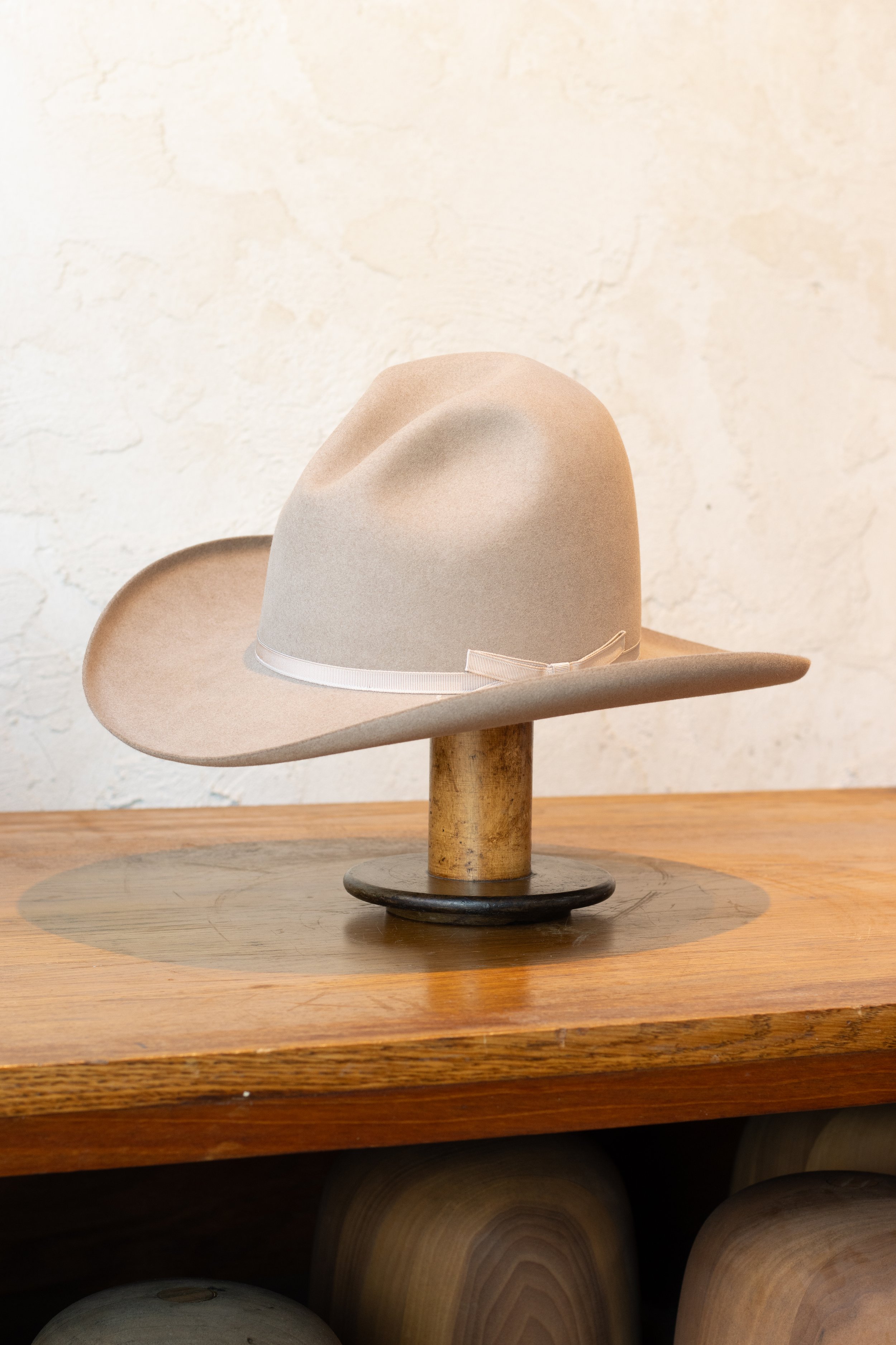 Cream-colored felt cowboy hat with a ribbon band, displayed on a wooden stand on a wooden surface.