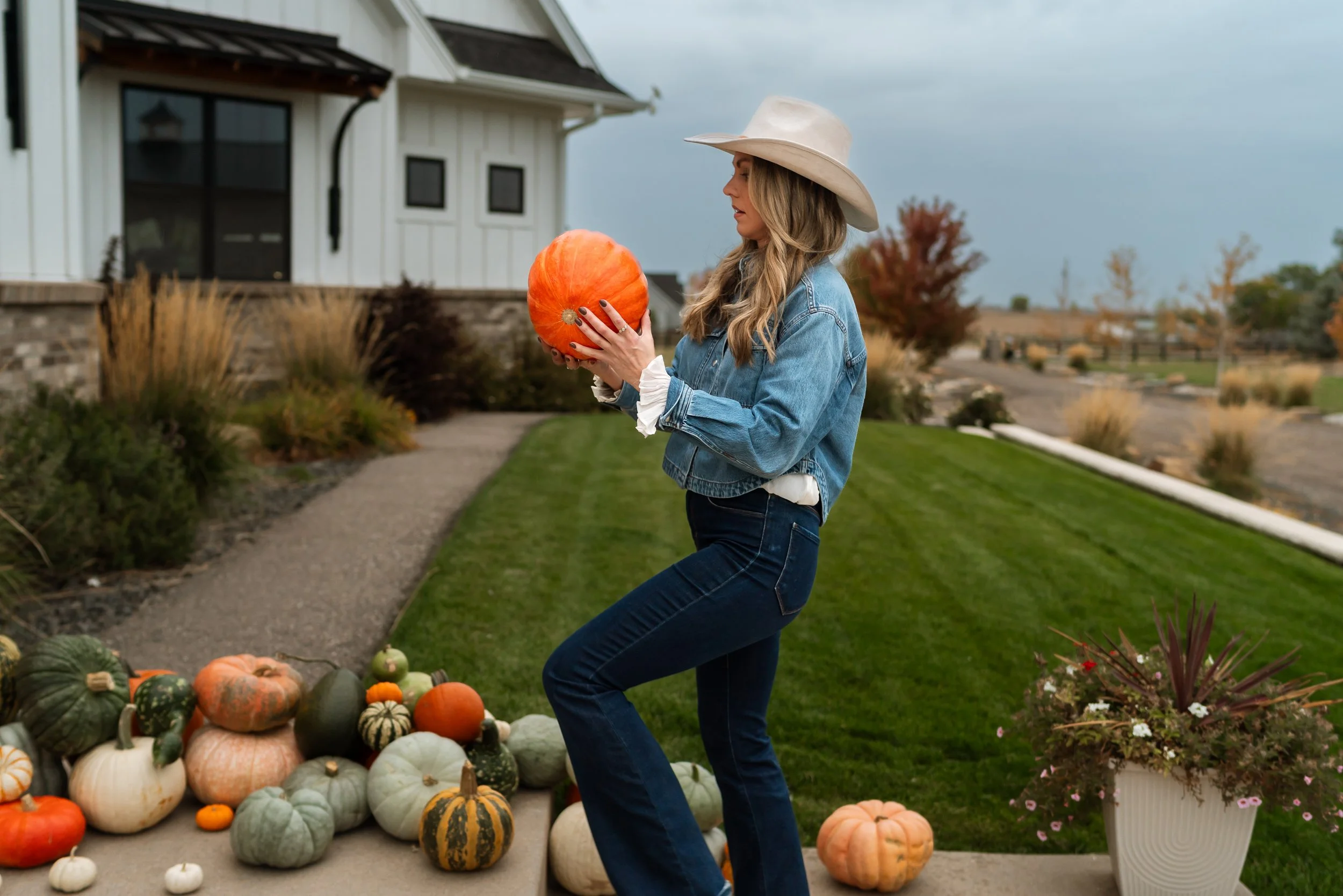 A woman in a cowboy hat and denim jacket holding a pumpkin while standing on a sidewalk decorated with pumpkins, gourds, and fall flowers outside a white house with a yard and trees.