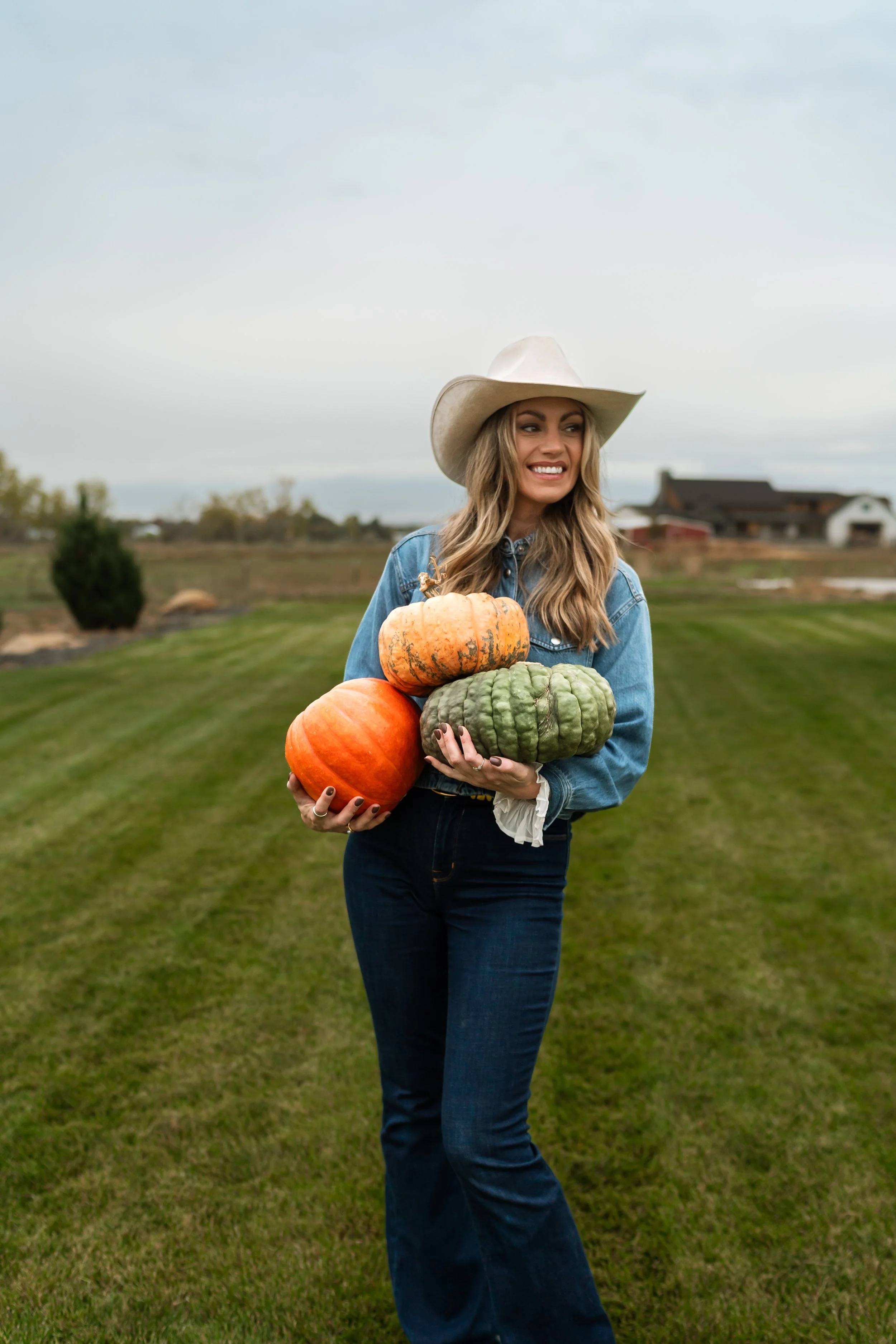 A woman with long wavy hair wearing a cowboy hat and denim jacket, holding a variety of colorful pumpkins in an outdoor rural setting with grass and farm buildings in the background.