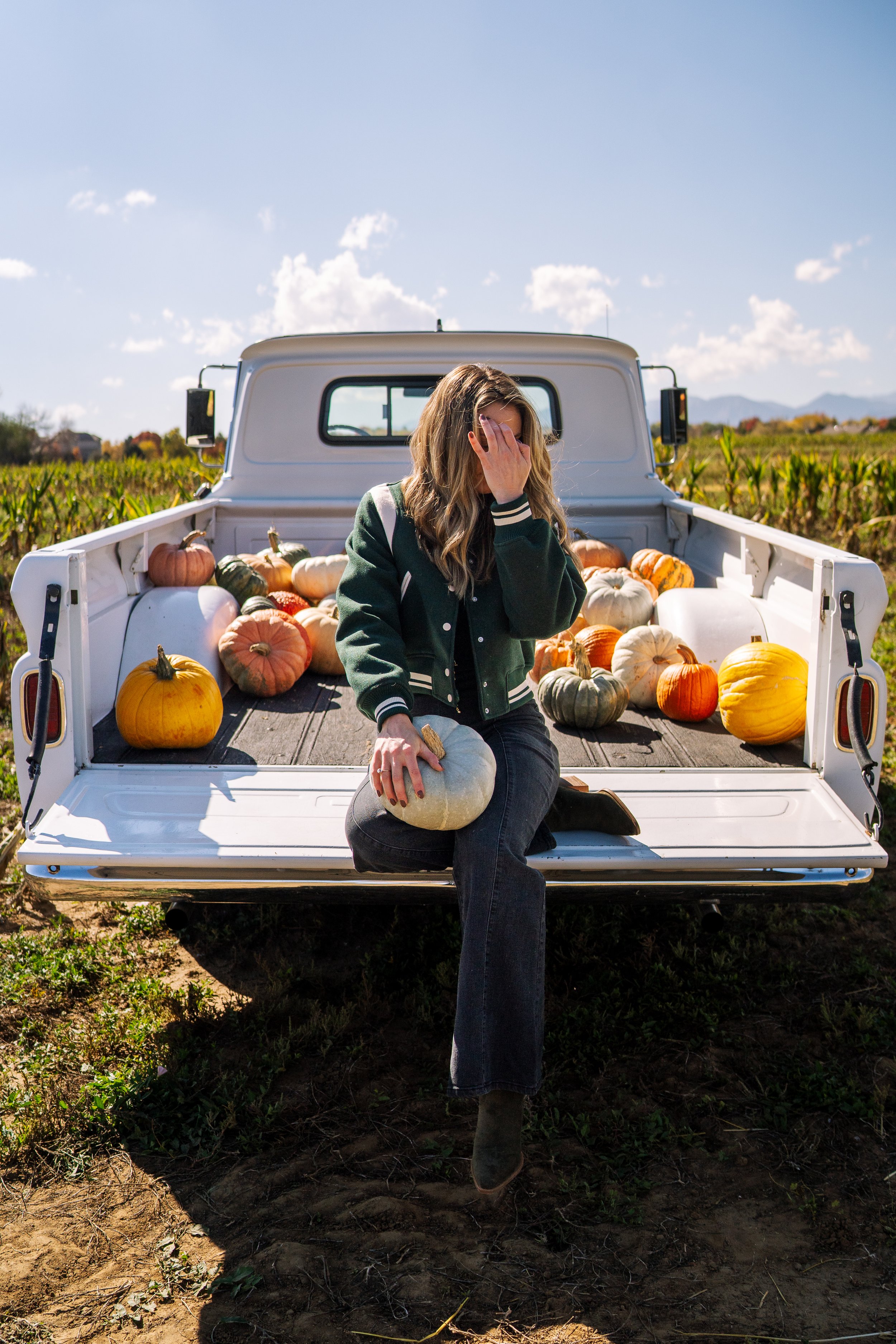 A woman sits on the tailgate of a pickup truck filled with pumpkins, covering her face with one hand. The scene is set in a pumpkin field on a sunny day.