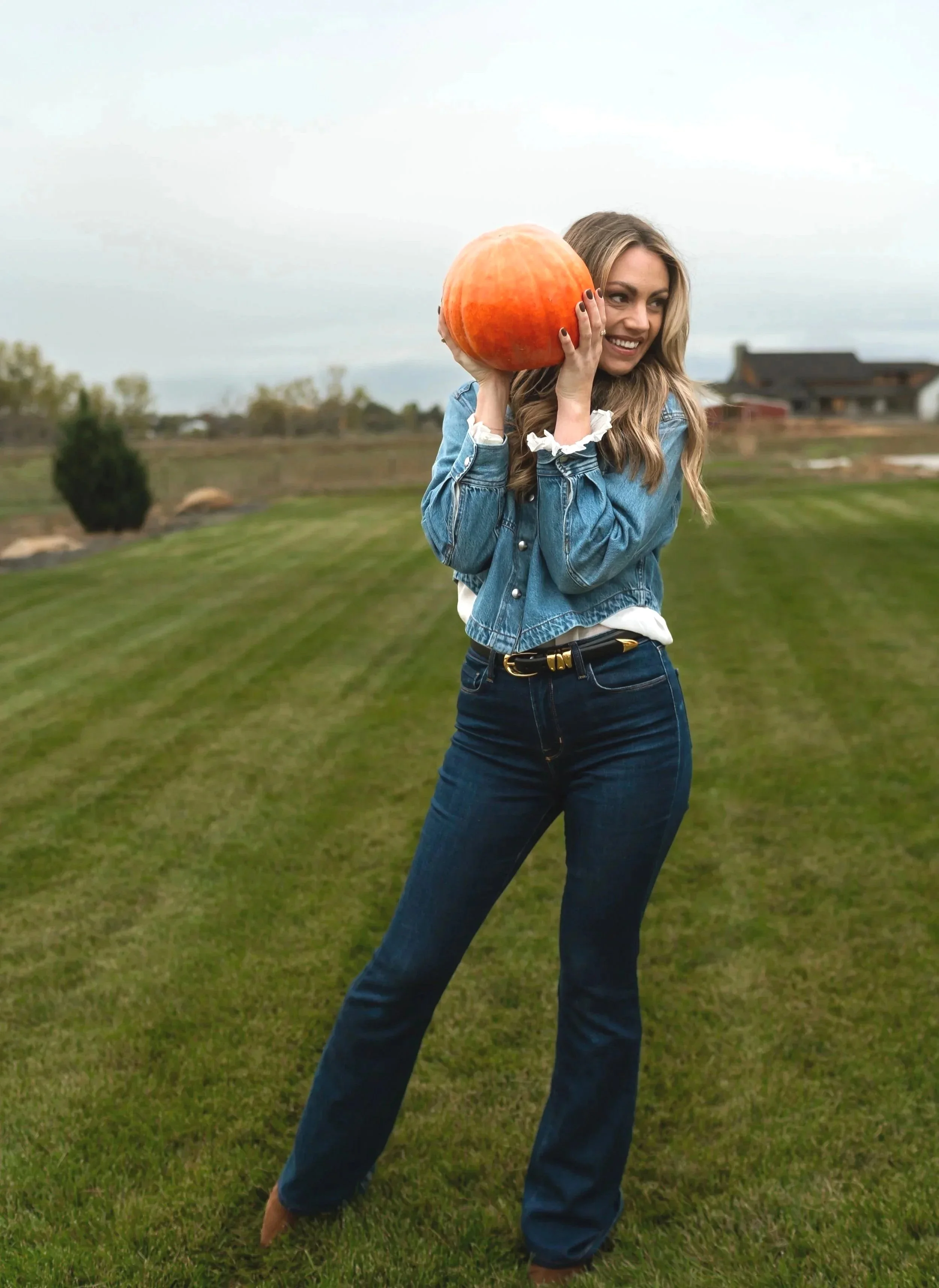 A woman with long wavy hair wearing a denim jacket and jeans, smiling while holding a large round orange pumpkin near her face outdoors on a grassy field with a rural house in the background.