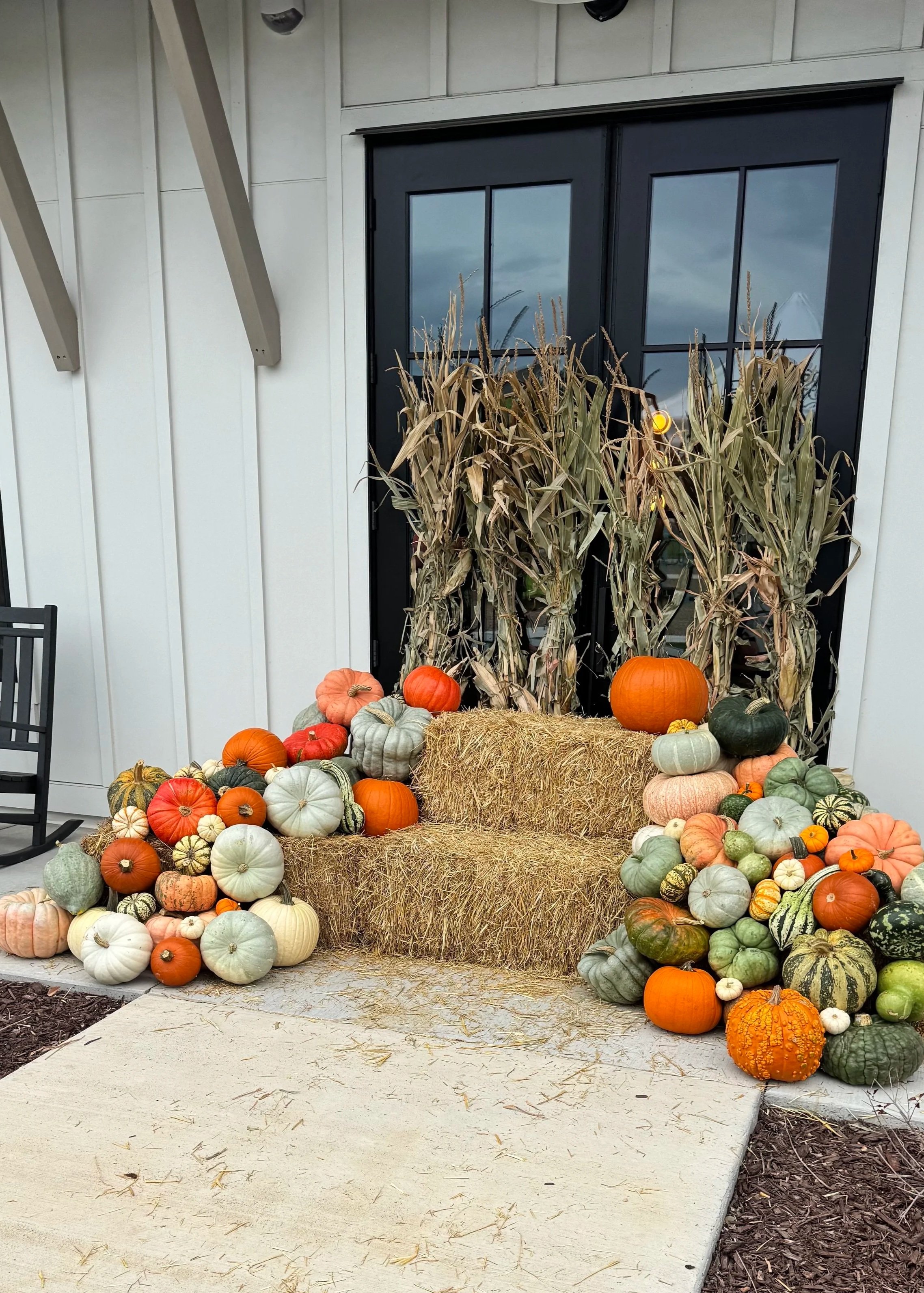 Fall harvest display on a porch with pumpkins, squash, gourds, hay bales, dried corn stalks, and a black-framed glass door