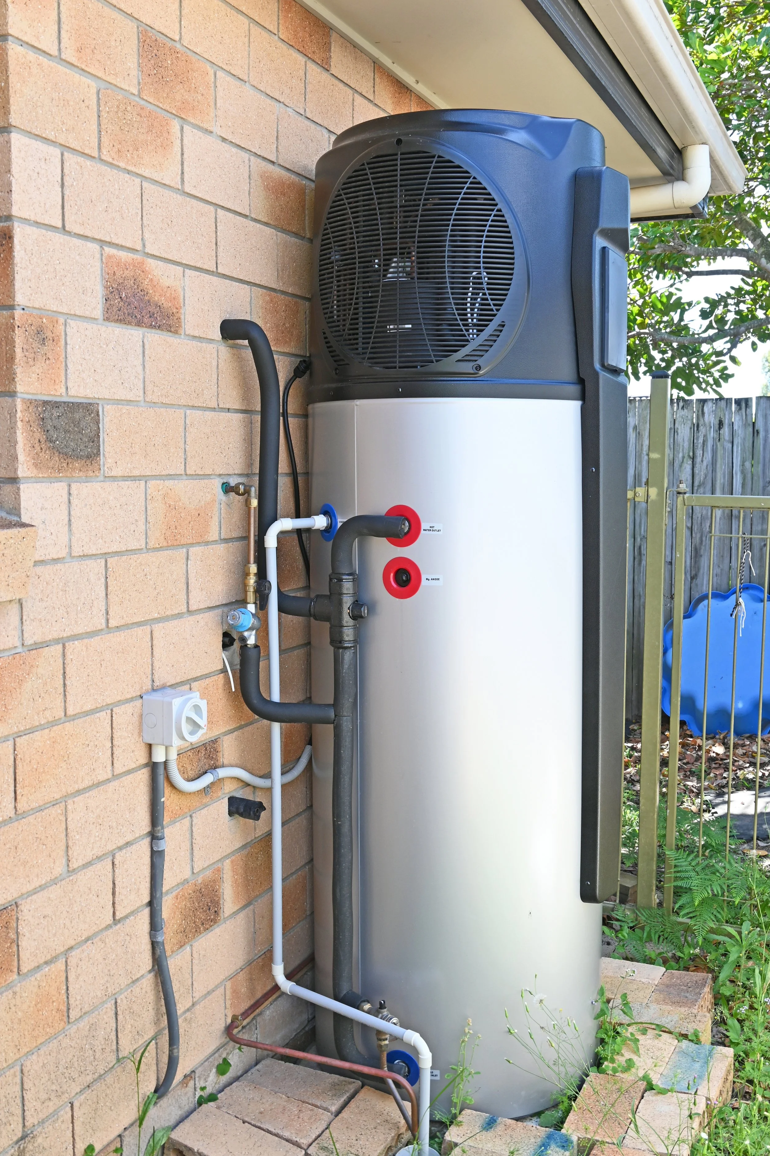 An outdoor air conditioning or heat pump unit installed next to a brick wall, with various pipes and electrical components connected.