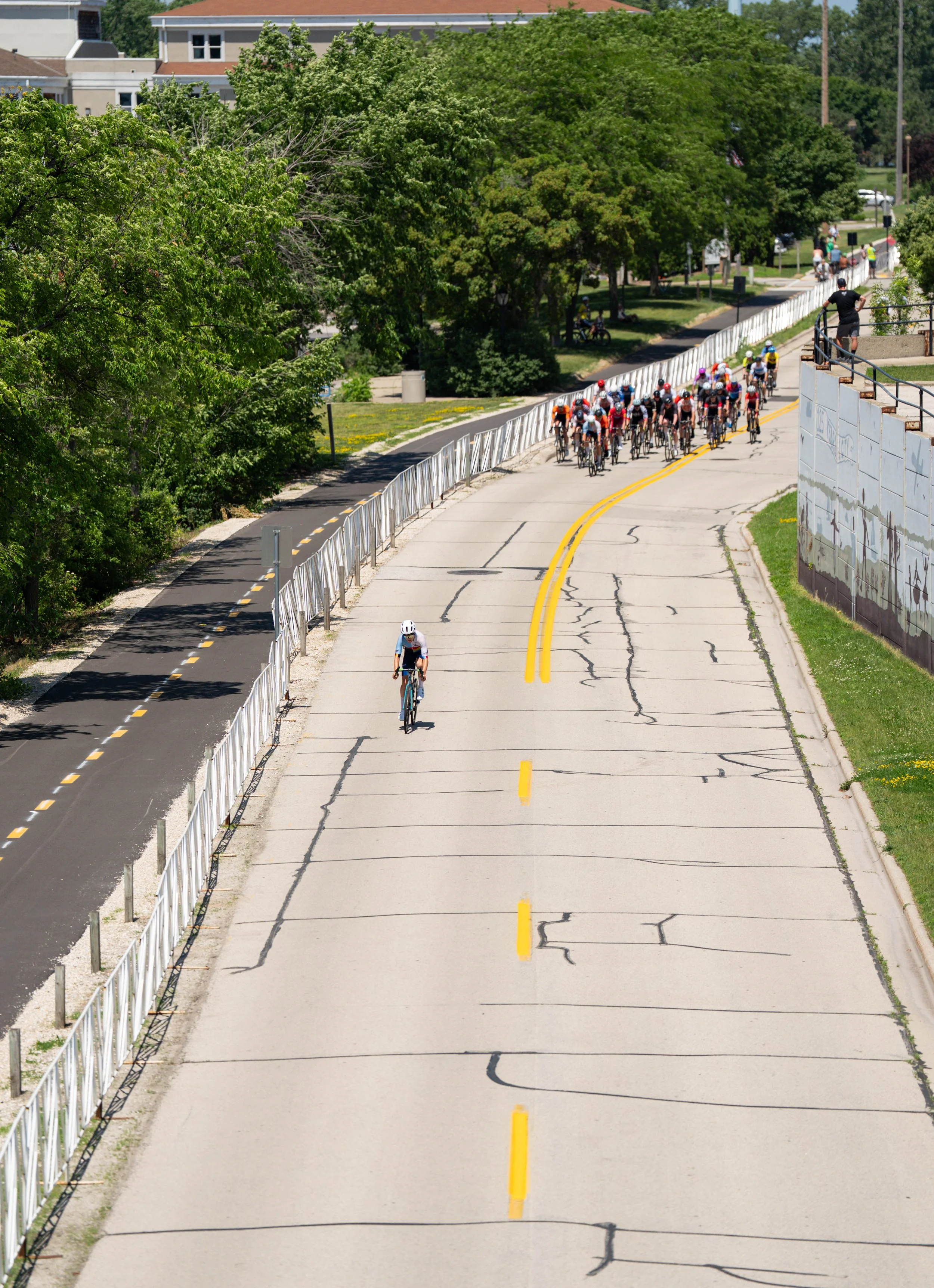 A group of cyclists riding on a cracked and weathered concrete road, with one cyclist ahead and the rest following behind, surrounded by green trees and city buildings in the background.