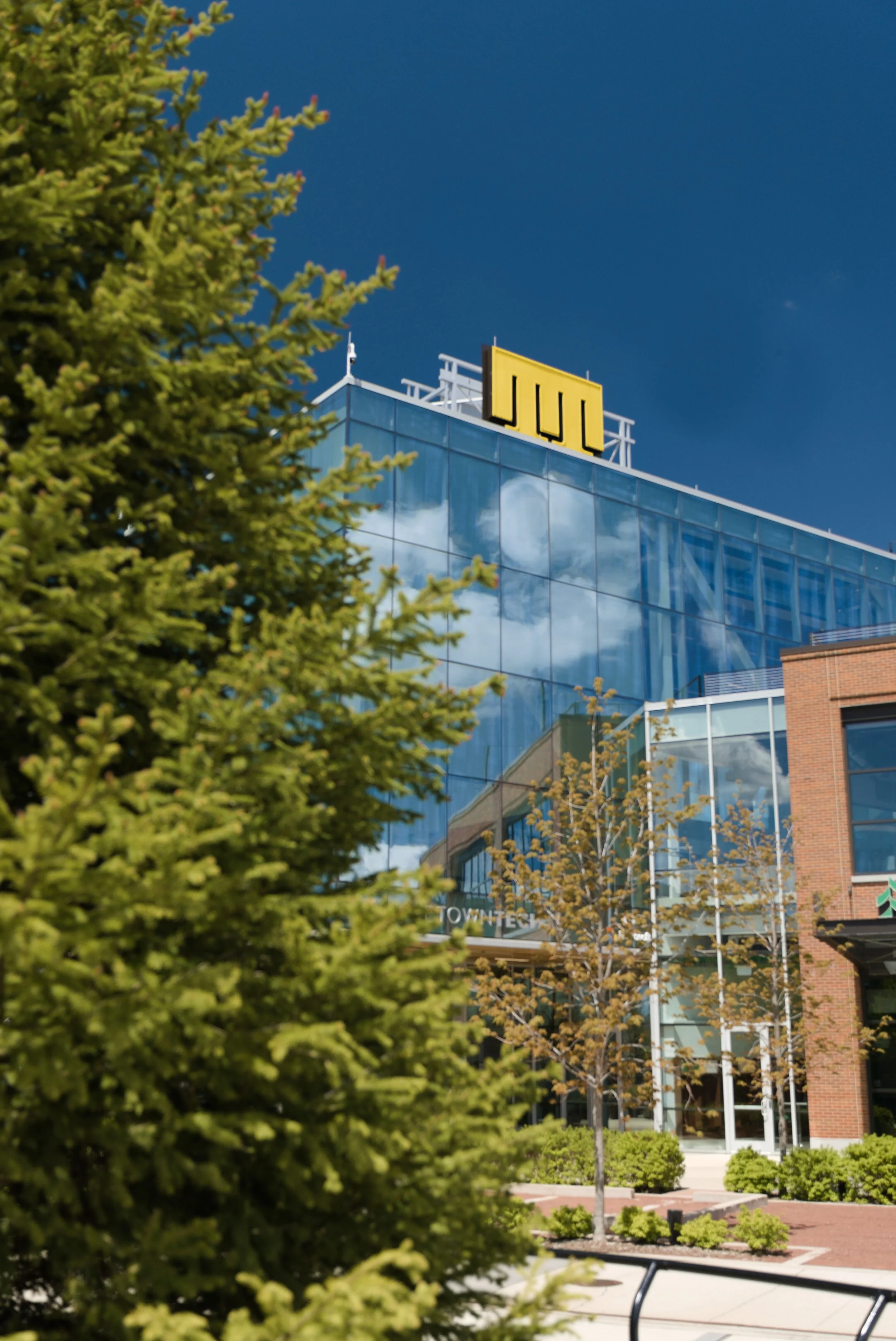 A modern glass building with a yellow sign on the roof that reads 'JLL'. In the foreground, there are trees with green leaves and a sidewalk, with a partly cloudy blue sky reflected in the glass.