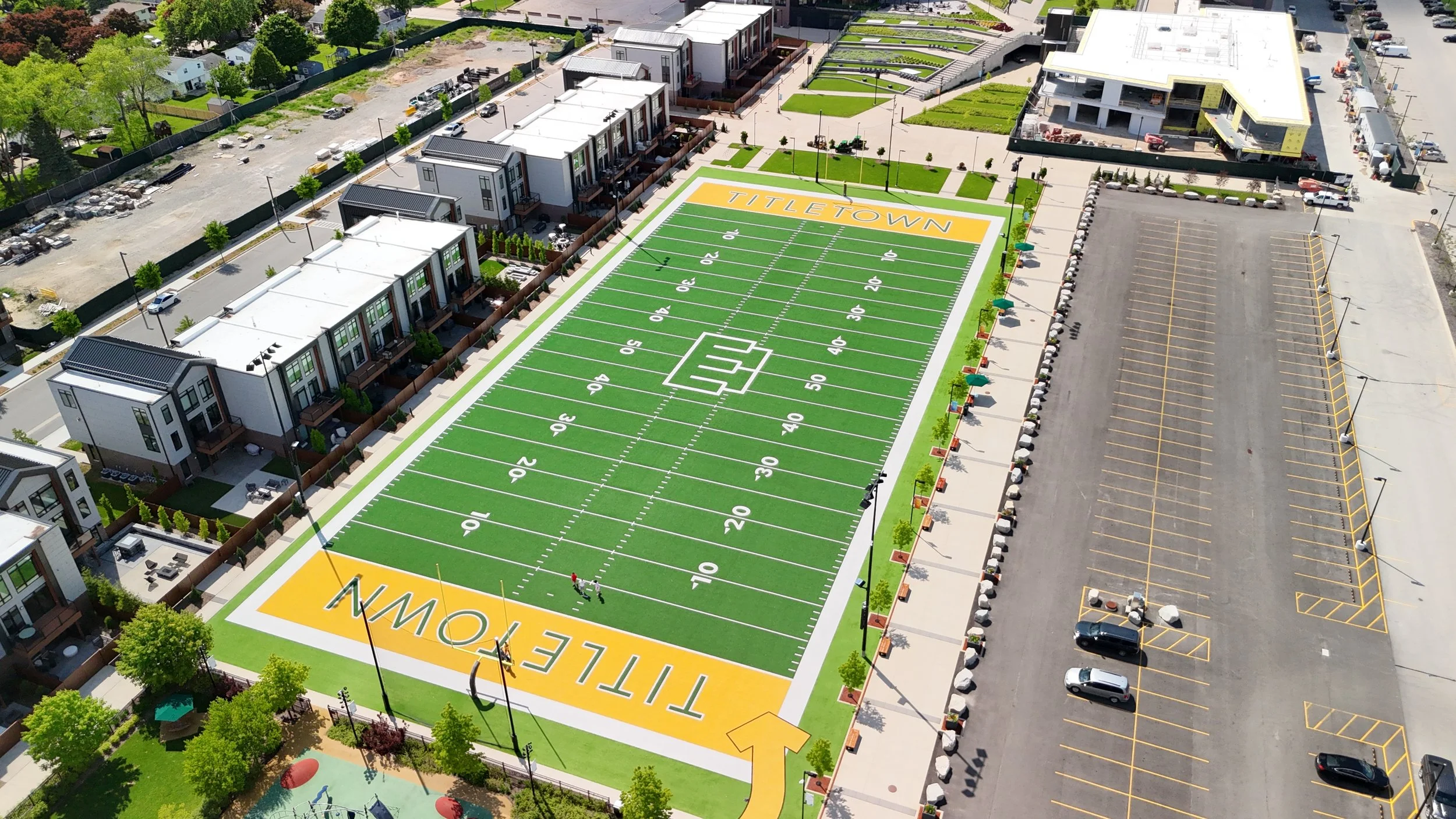 Aerial view of a football field with green turf marked with yard lines from 10 to 50, the word 'TITLETOWN' at one end zone, and a large logo at the center. The field is surrounded by a sidewalk, trees, modern residential buildings, and a large empty parking lot with painted parking spaces and a few vehicles.