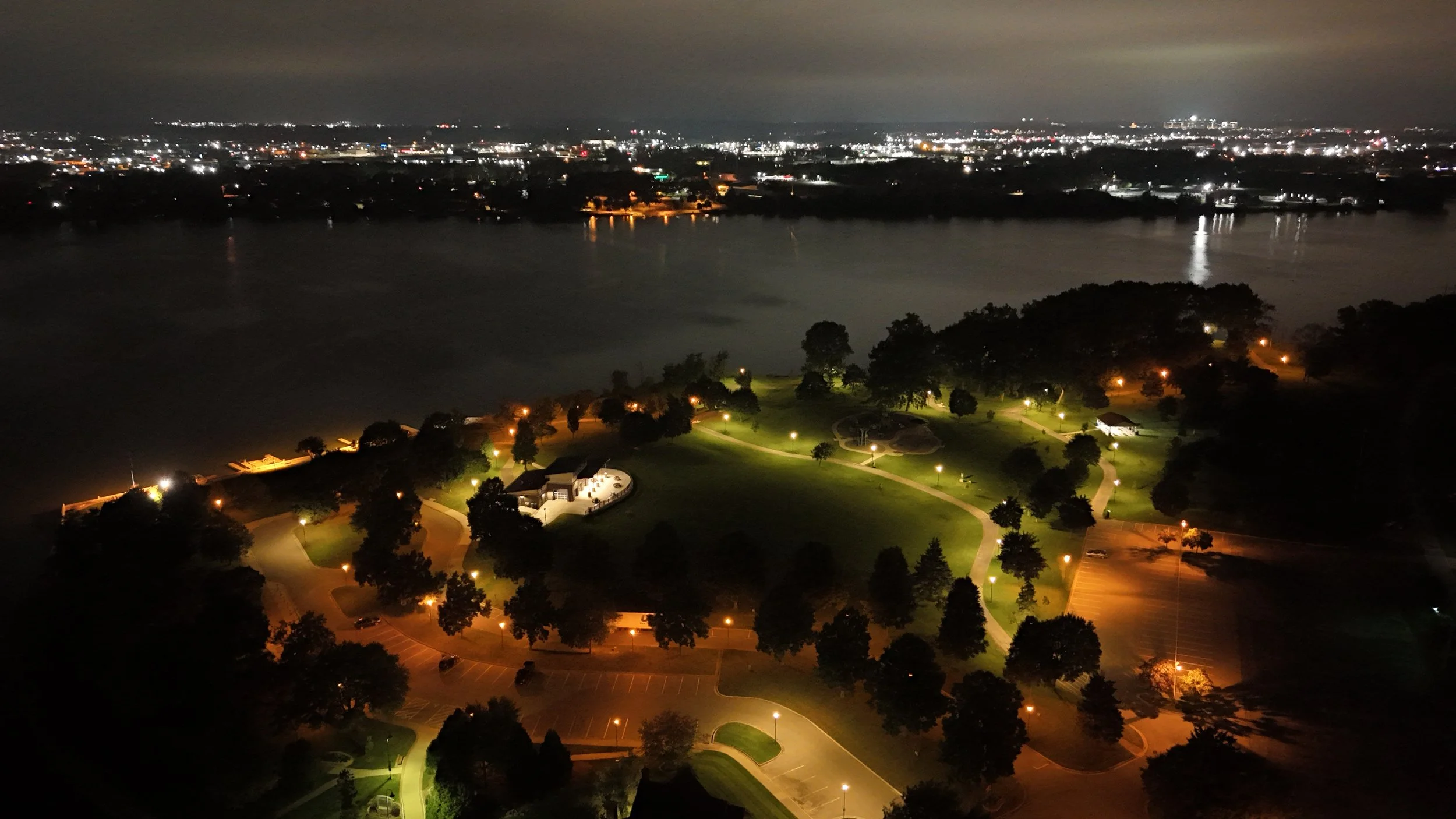 Night view of a park by a river with illuminated pathways and trees, with city lights visible across the water in the background.