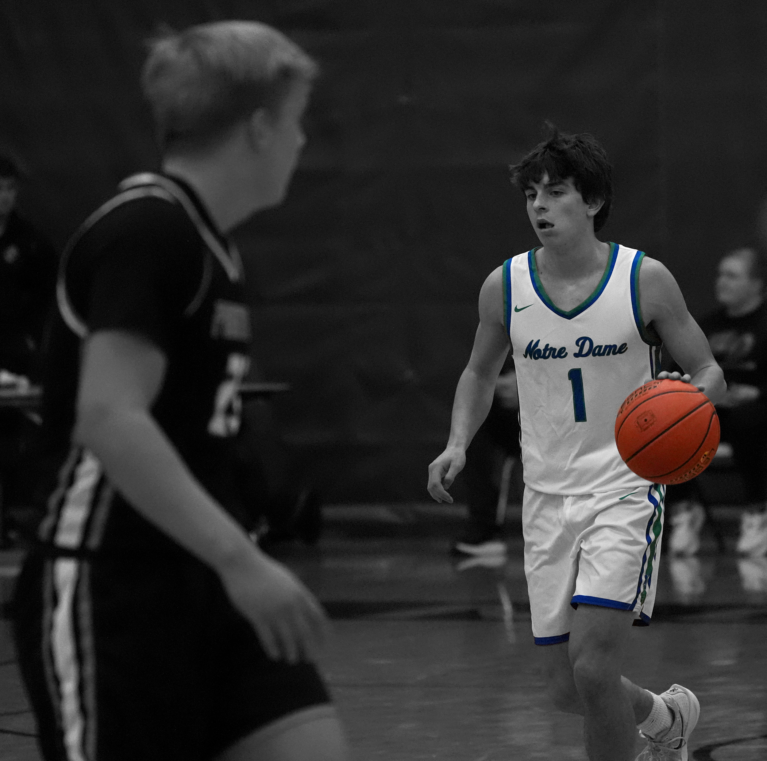 A young basketball player wearing a Notre Dame jersey with the number 1 dribbling a basketball on an indoor court, with another player in the foreground and a woman sitting in the background.