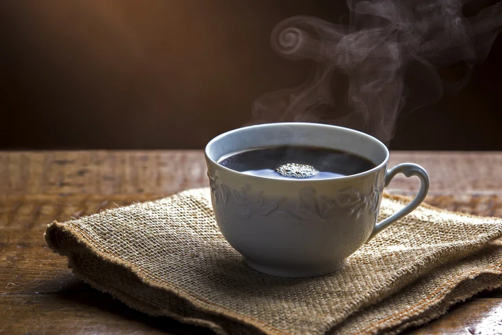 A steaming cup of black coffee on a piece of burlap cloth on a wooden surface.