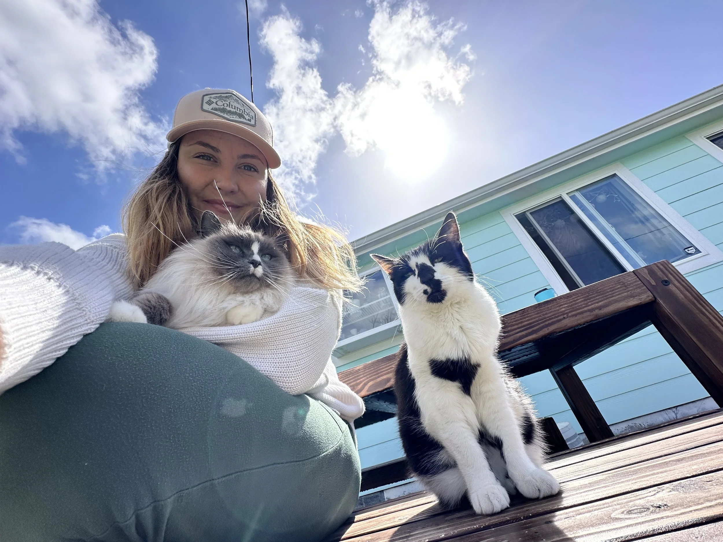 A woman with long hair wearing a beige baseball cap and white sweater, holding a Siamese cat while another black and white cat sits on a wooden deck beside her, in front of a blue house under a partly cloudy sky.