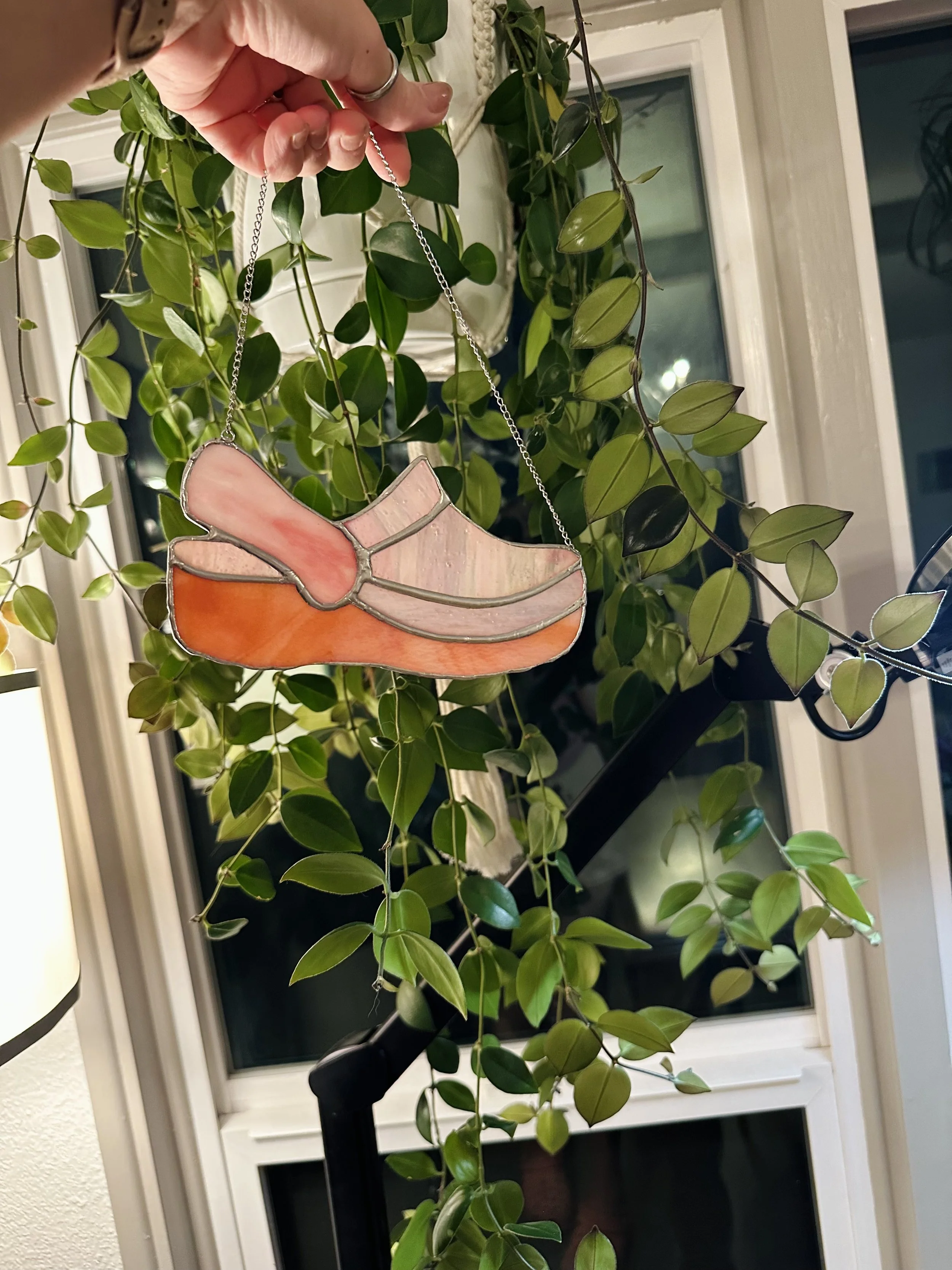 A hand holding a stained glass ornament in the shape of an orange and pink shoe hanging from a silver chain, with a green leafy houseplant in the background.