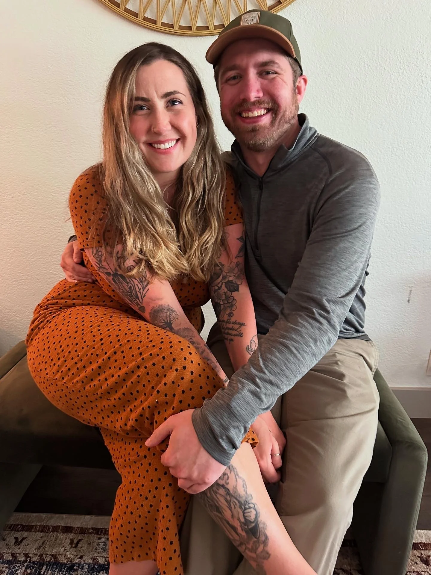 A smiling woman with long blonde hair, tattoos on her arms and leg, wearing an orange polka dot dress, sitting on a man's lap, hugging him. The man has a beard, mustache, short hair, wearing a green cap, gray zip-up shirt, and beige pants. They are in a room with a plain wall and a decorative wall hanging behind them.