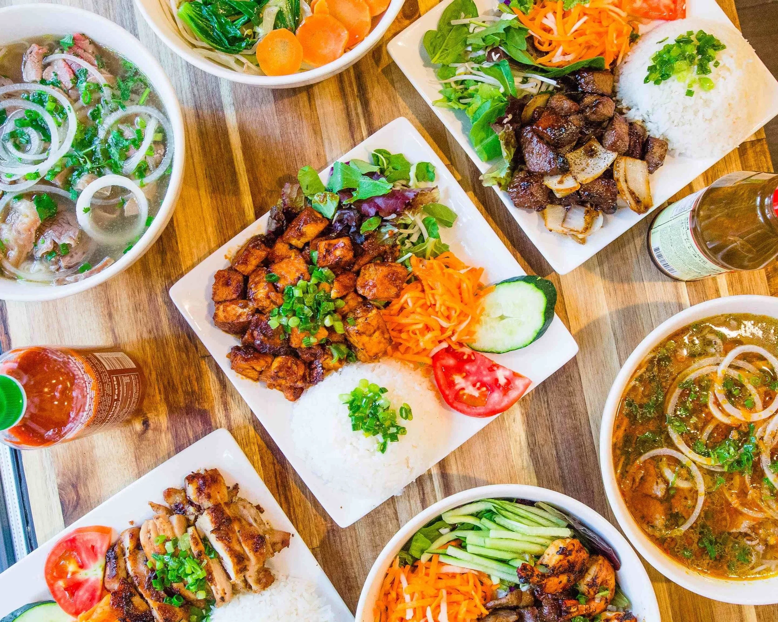 Assorted Vietnamese dishes on a wooden table, including bowls of noodle soup, rice plates with grilled meats and vegetables, and side salads with fresh herbs and vegetables.
