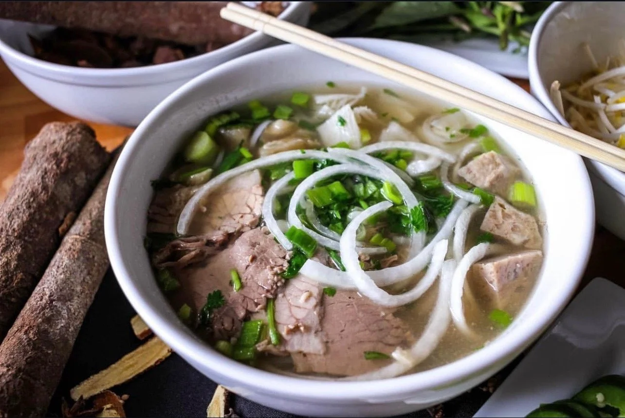 Bowl of Vietnamese pho with slices of beef, onions, and chopped green onions, accompanied by chopsticks, on a wooden table with side dishes and herbs.