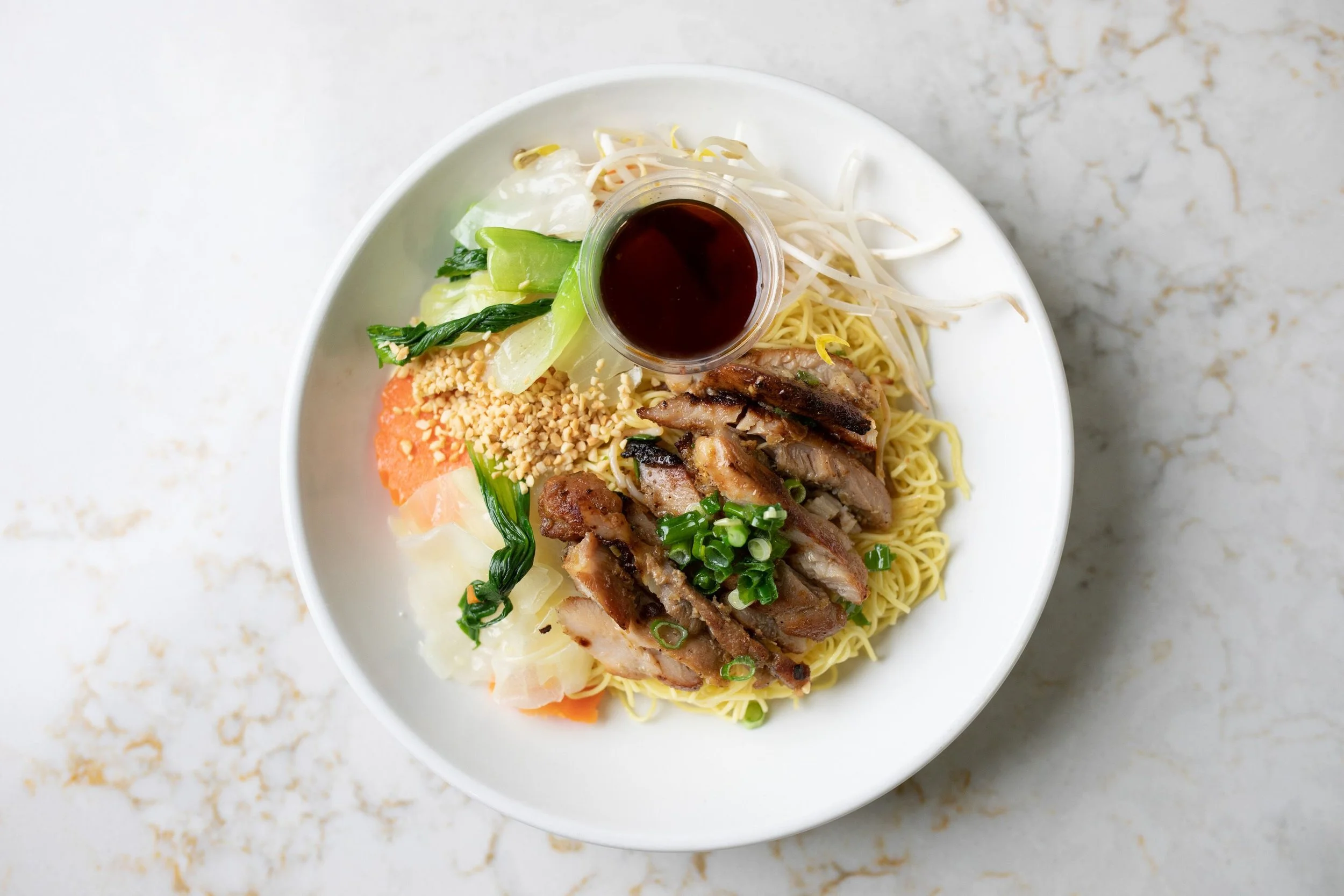 Bowl of Asian noodle salad with grilled chicken, fresh vegetables, chopped peanuts, bean sprouts, and a small container of soy sauce on top.