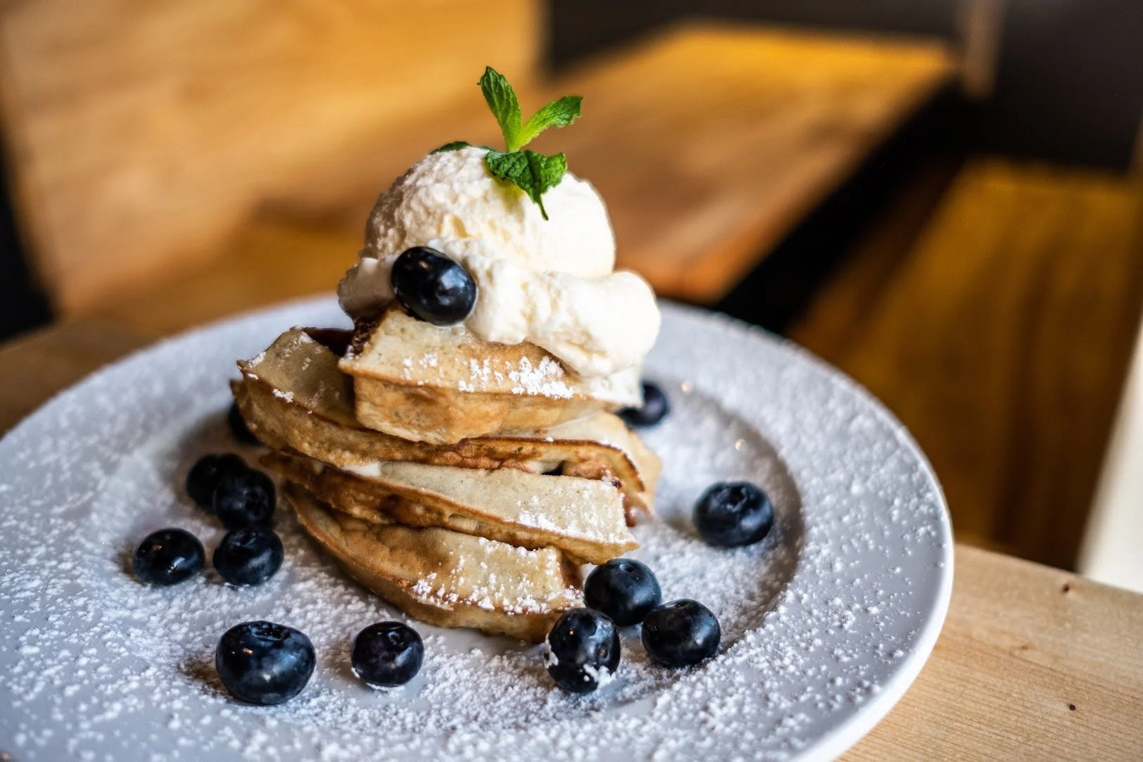 Stack of waffles topped with whipped cream, a scoop of vanilla ice cream, blueberries, a sprig of mint, and powdered sugar on a white plate.