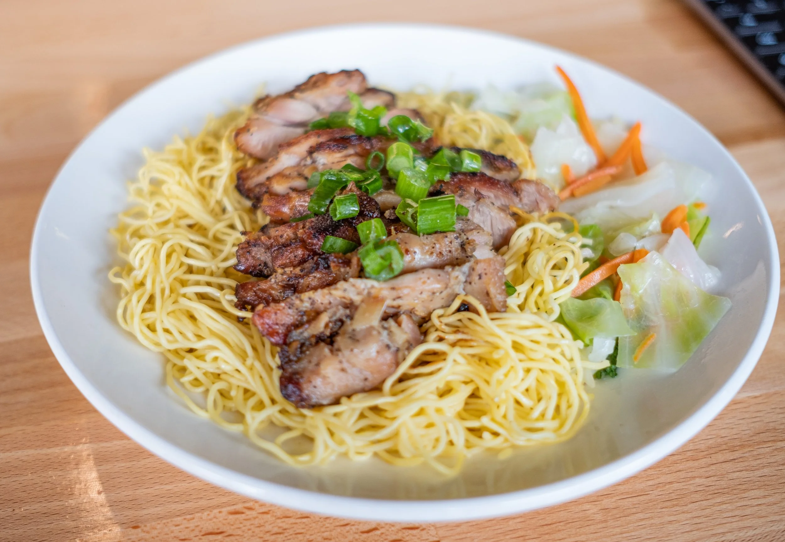 A white bowl containing noodles topped with grilled chicken slices garnished with chopped green onions, alongside mixed vegetables including lettuce, carrots, and cabbage, on a wooden table.