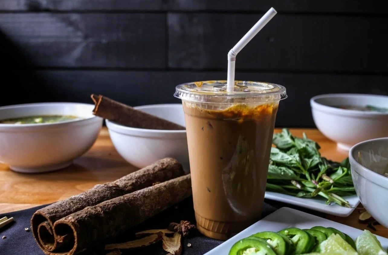 A plastic cup of iced coffee with a straw surrounded by bowls of herbs and vegetables, and cinnamon sticks on a wooden table.