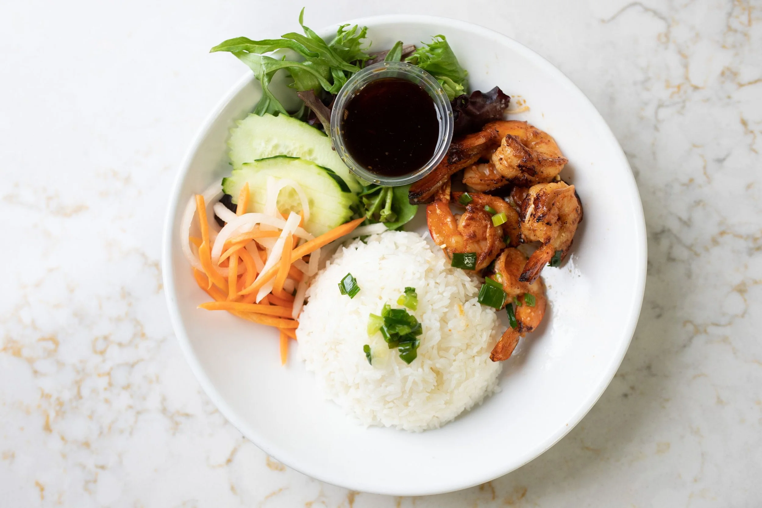 Plate of grilled shrimp with rice, fresh salad with cucumbers, carrots, mixed greens, and a small container of soy sauce on a white marble surface.