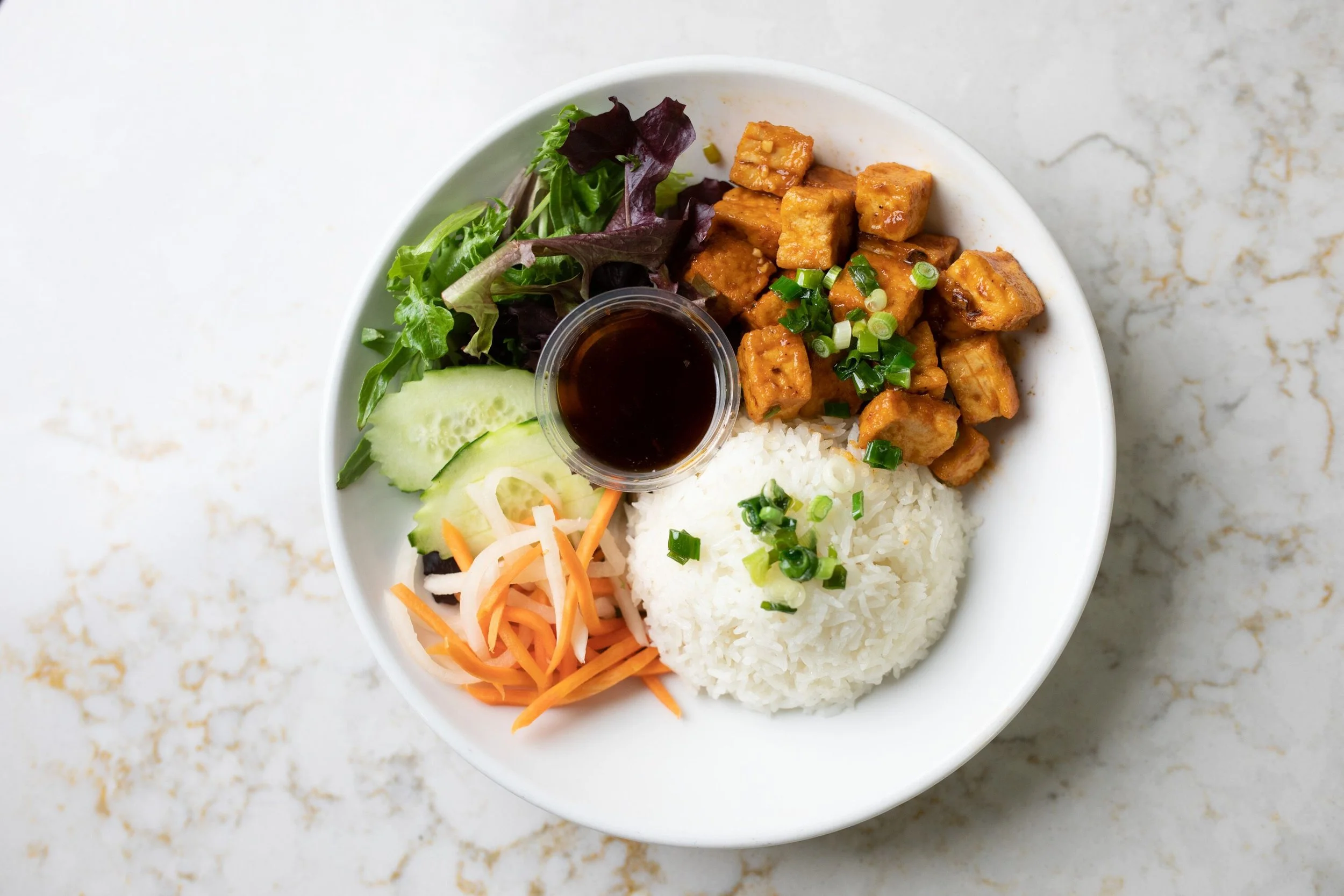 A white bowl with rice, tofu, mixed greens, cucumber, shredded carrots, and a small cup of soy sauce, served on a marble surface.