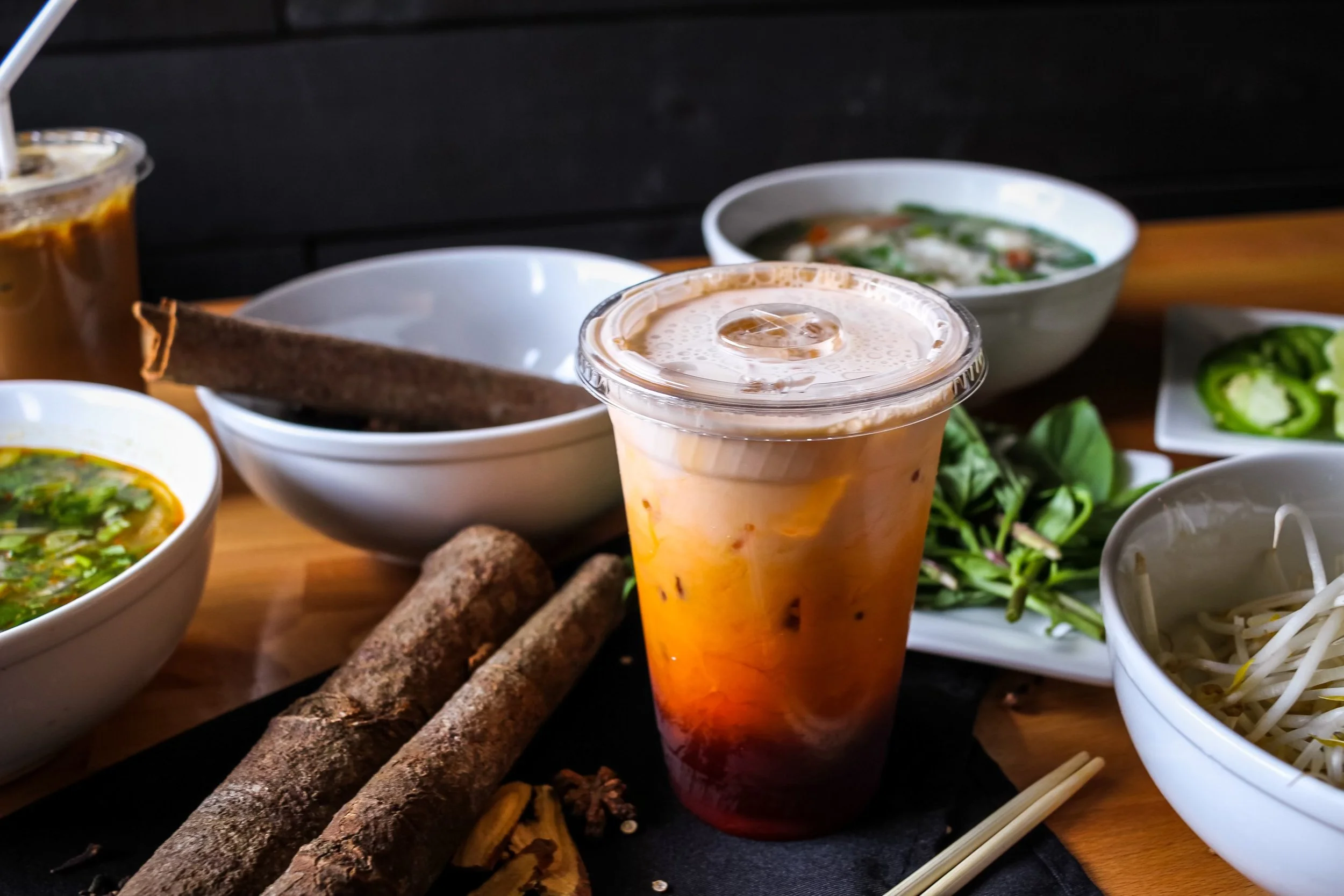 Assorted Vietnamese dishes with a cold drink, including soups, fresh herbs, and vegetables on a wooden table.