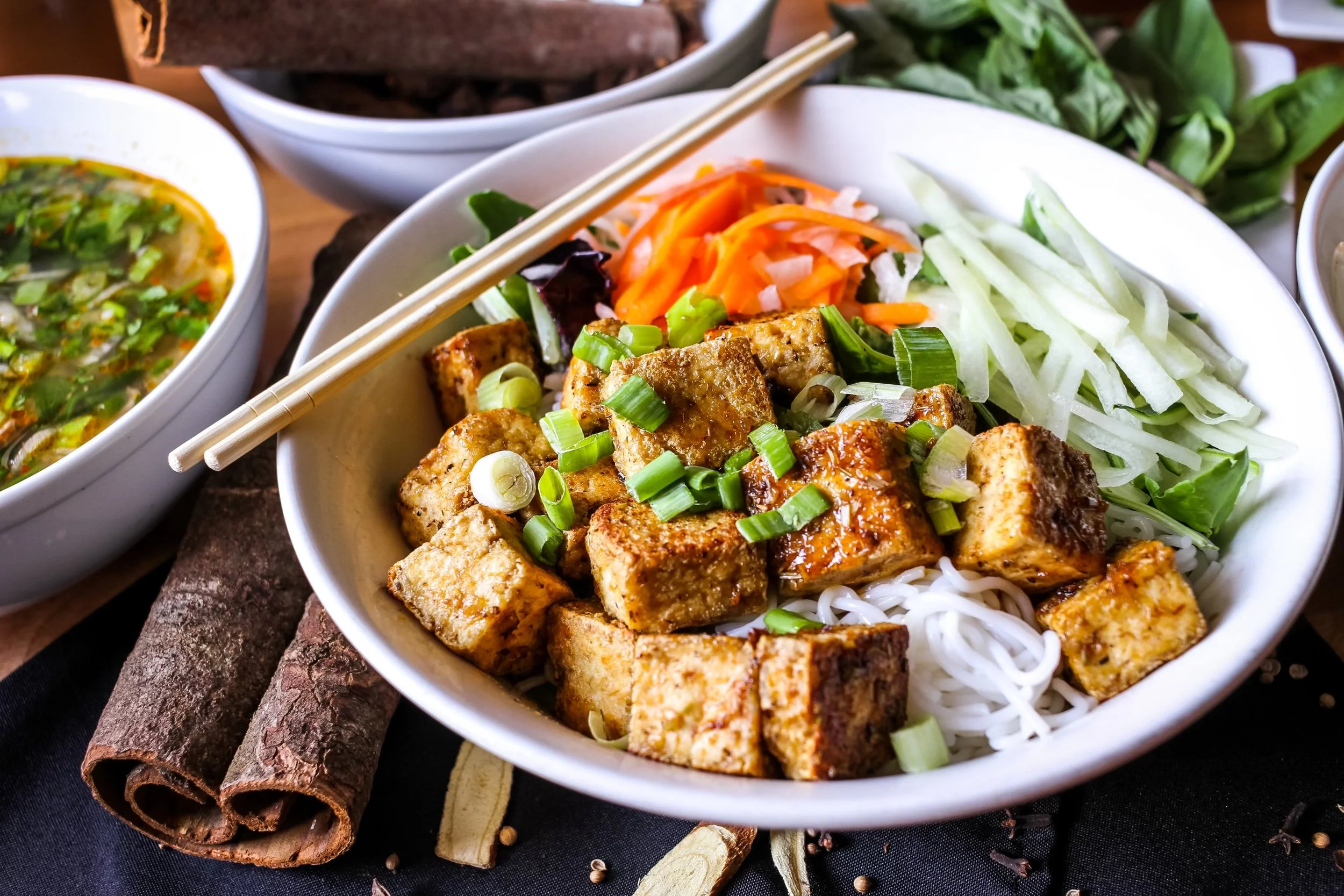 Bowl of tofu, rice noodles, and fresh vegetables with chopped green onions on top, paired with chopsticks, on a dark surface with cinnamon sticks and spice scattered around