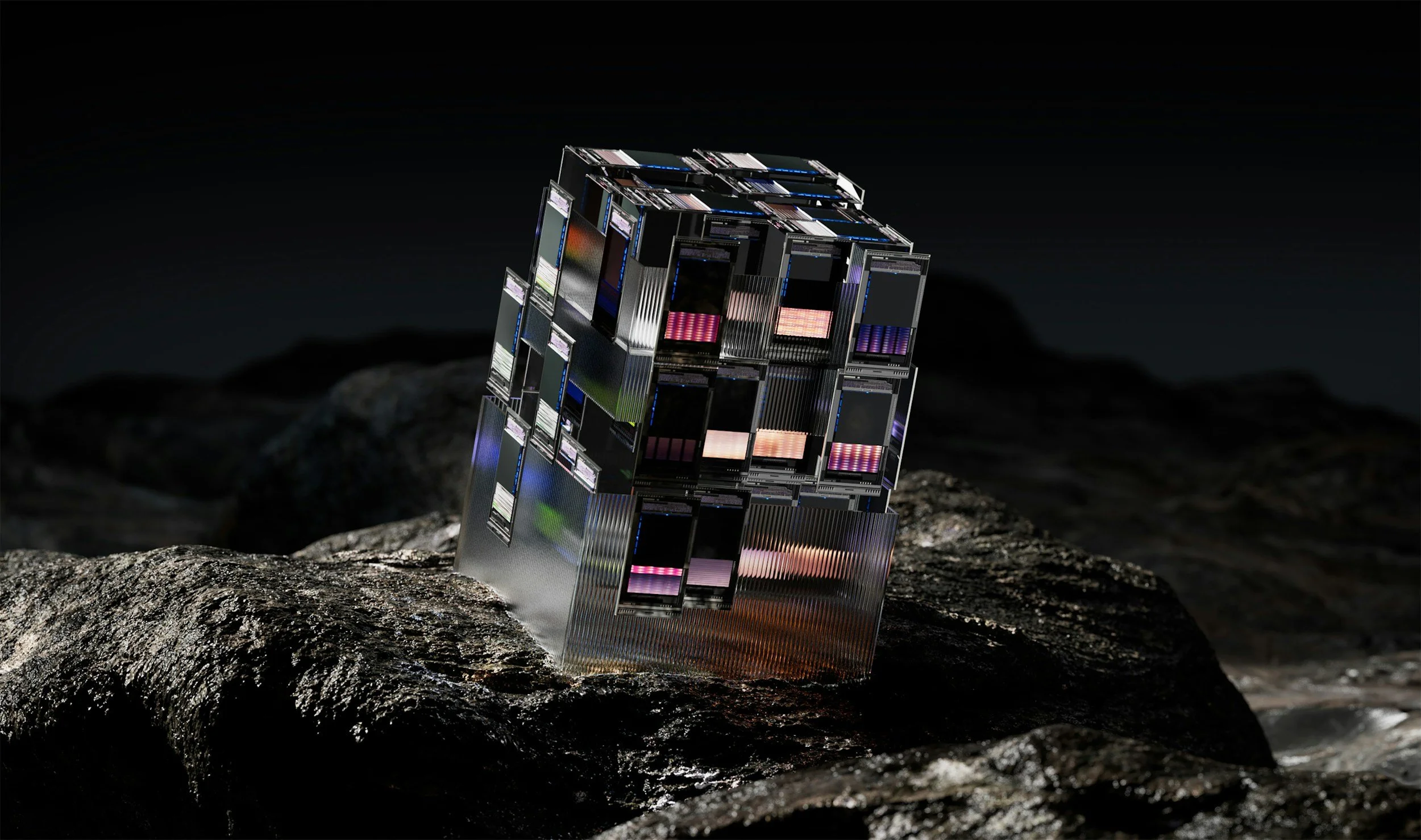 A metallic sculpture resembling a Rubik's Cube, composed of multiple computer screens displaying colorful digital patterns, sitting on a rough rock surface against a dark background.