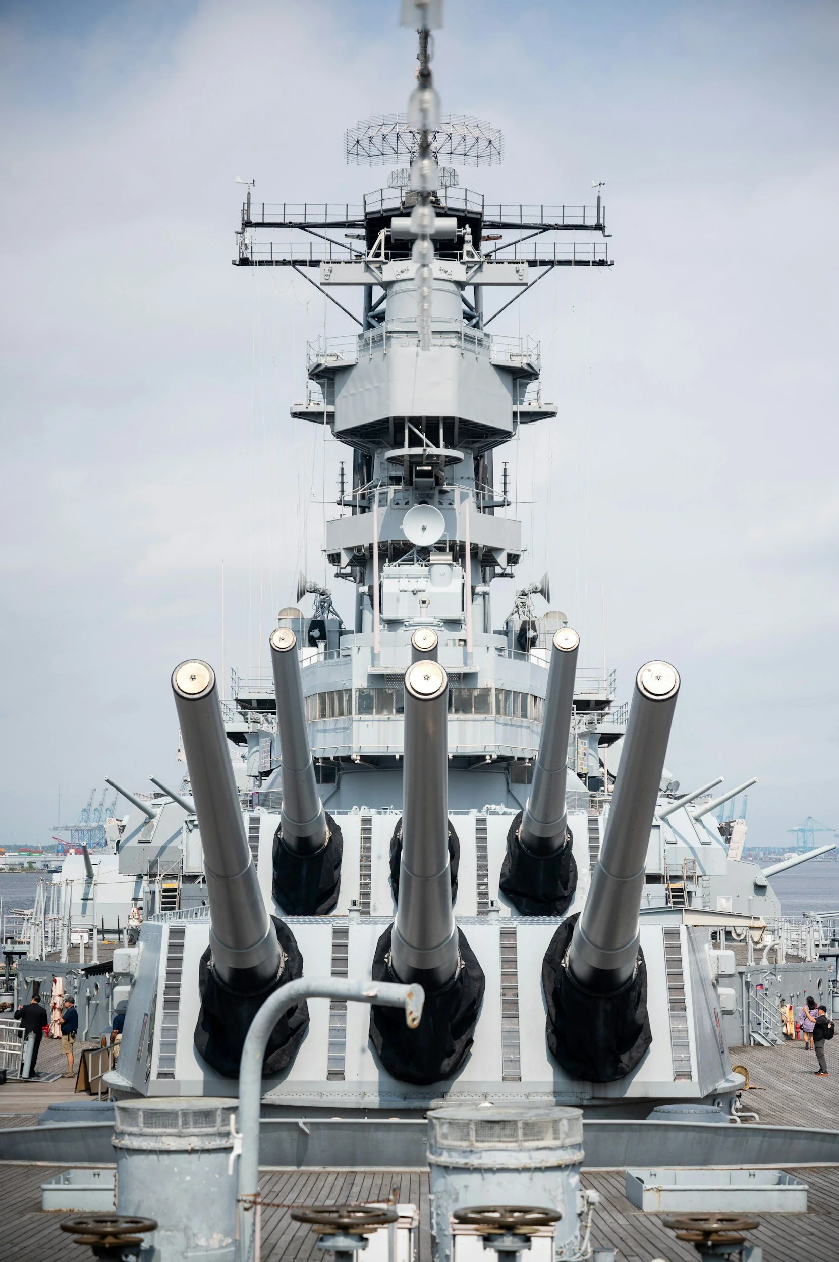Front view of a large military battleship with four large gun barrels pointing forward, docked at a port with people walking nearby and cranes in the background.