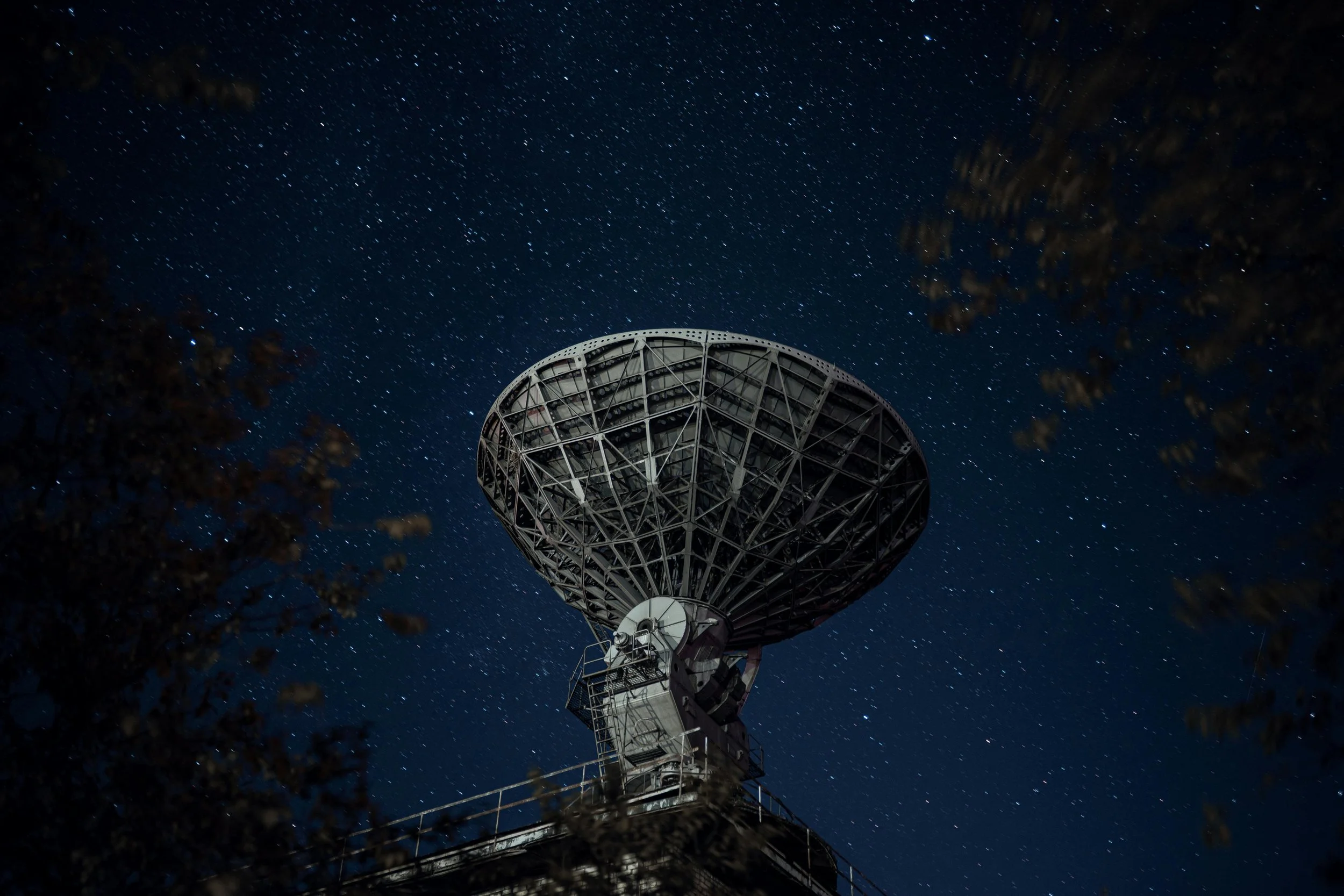Satellite dish under a starry night sky with branches in the foreground.
