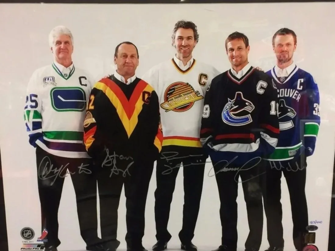 Group photo of former captains of the Vancouver Canucks wearing Vancouver Canucks hockey jerseys with signatures, standing in a row.