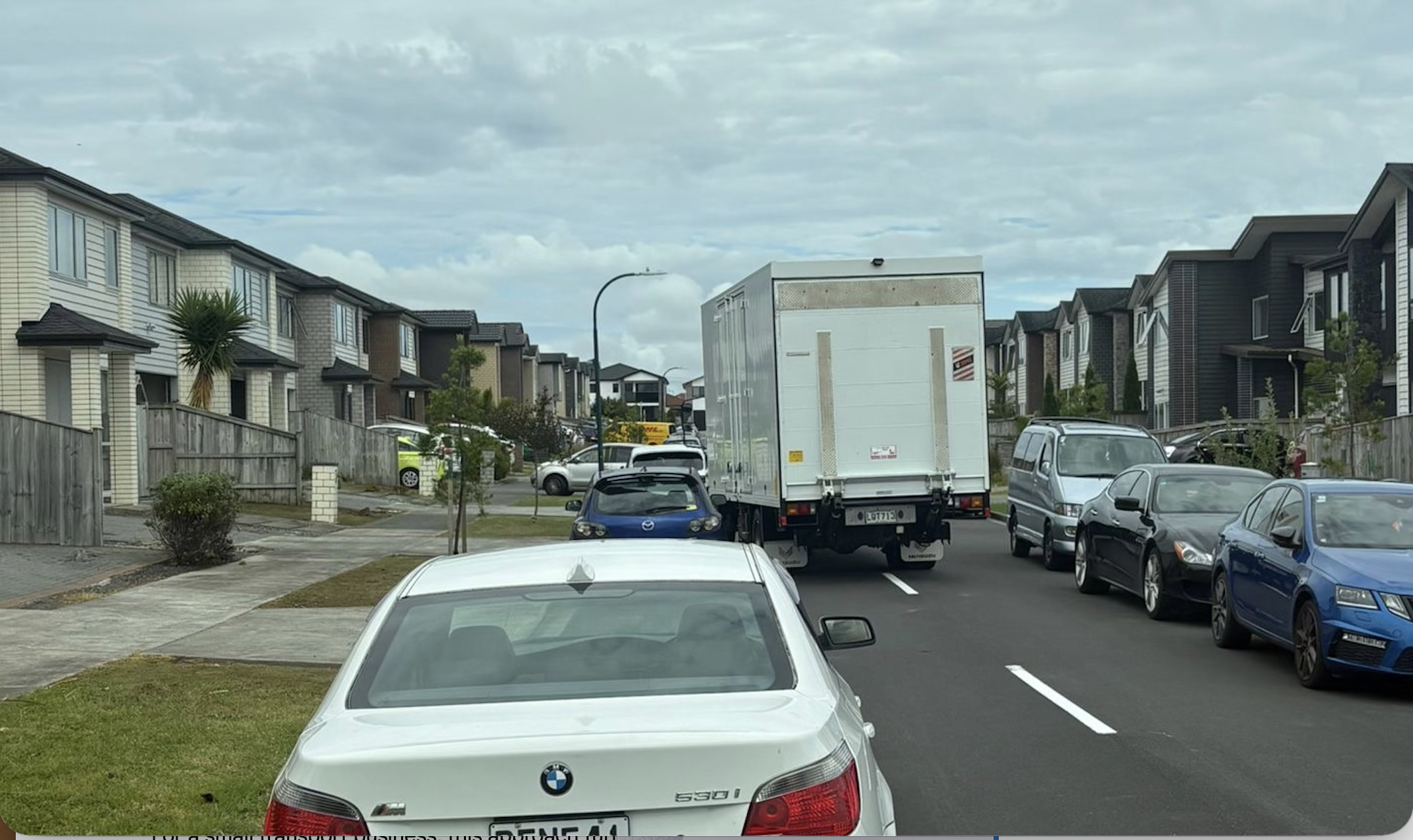 Residential street scene with parked cars, a white delivery truck blocking the road, and houses on both sides under a cloudy sky.