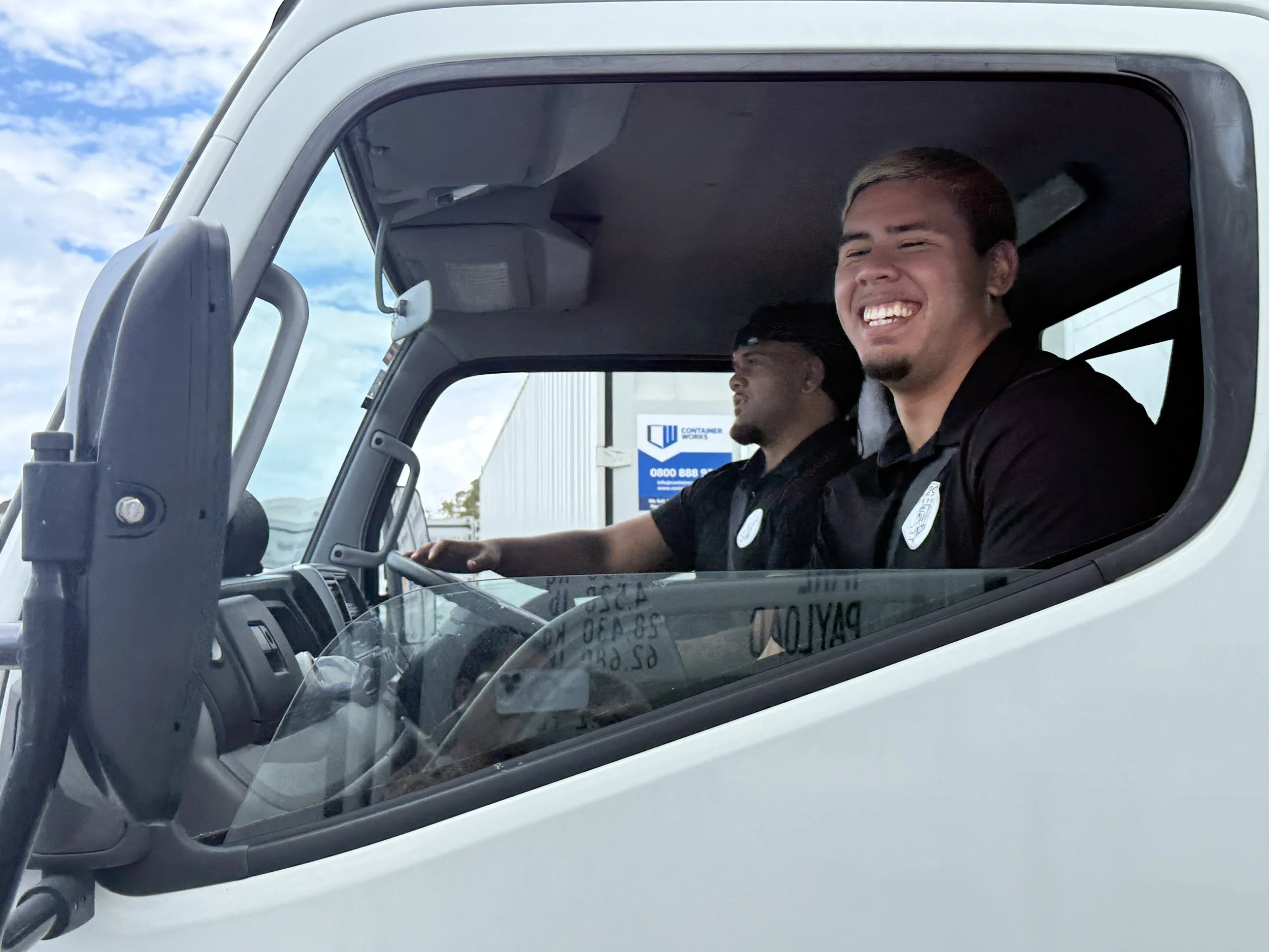 Two men in black uniforms sitting inside a white truck, smiling happily, with a blue sky and clouds visible through the window.