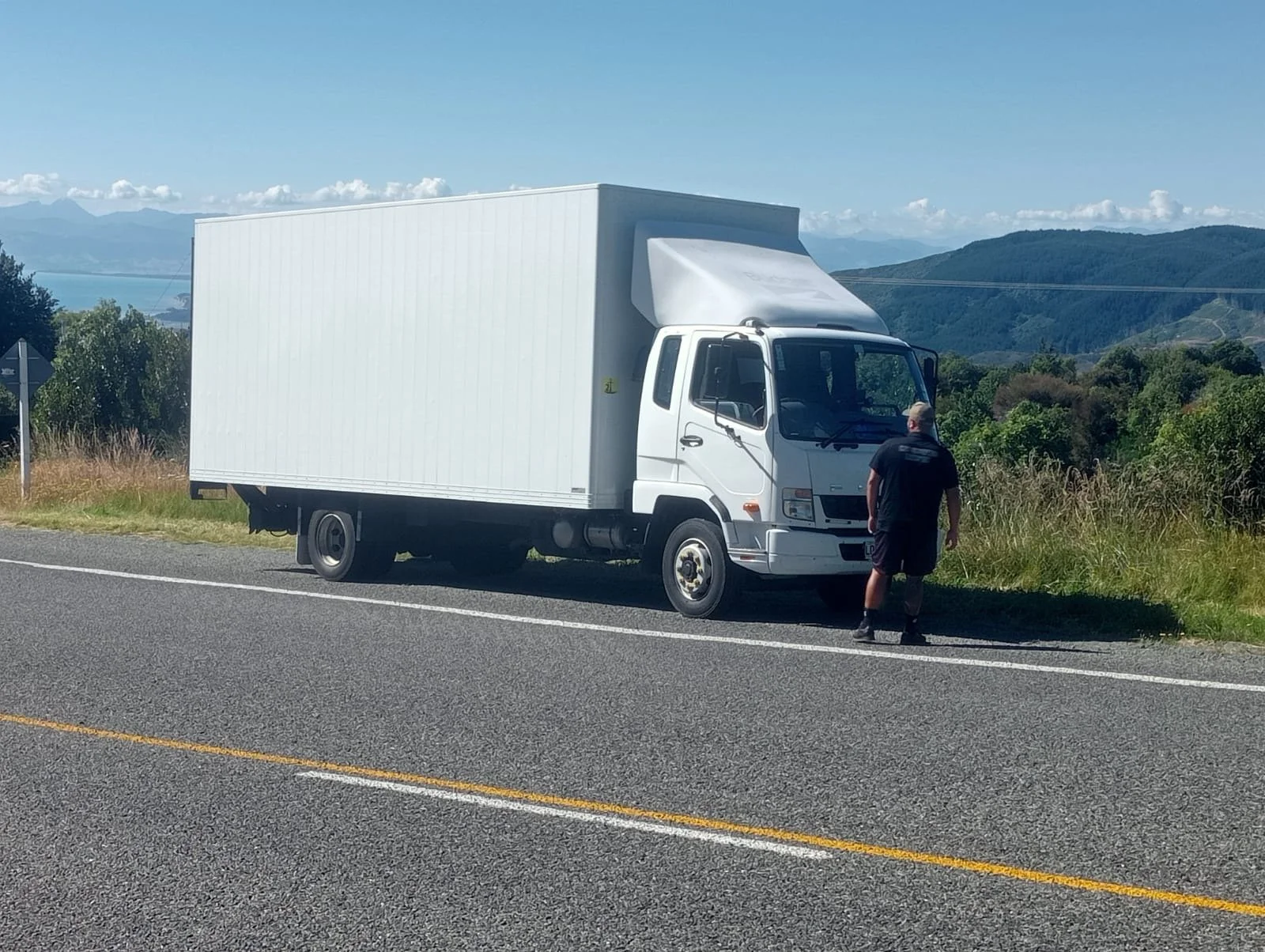 A white moving truck parked on the side of a two-lane road, with a man standing next to it, against a background of green trees, mountains, and a partly cloudy sky.