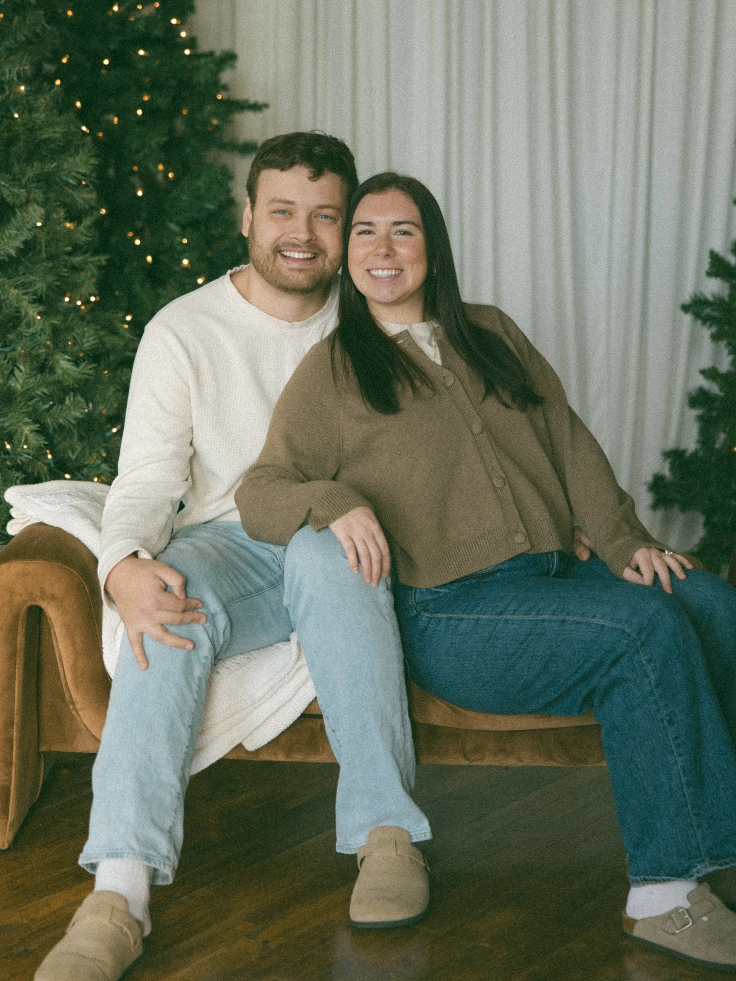A smiling couple sitting on a vintage sofa in front of decorated Christmas trees with lights, in a cozy room with white curtains.