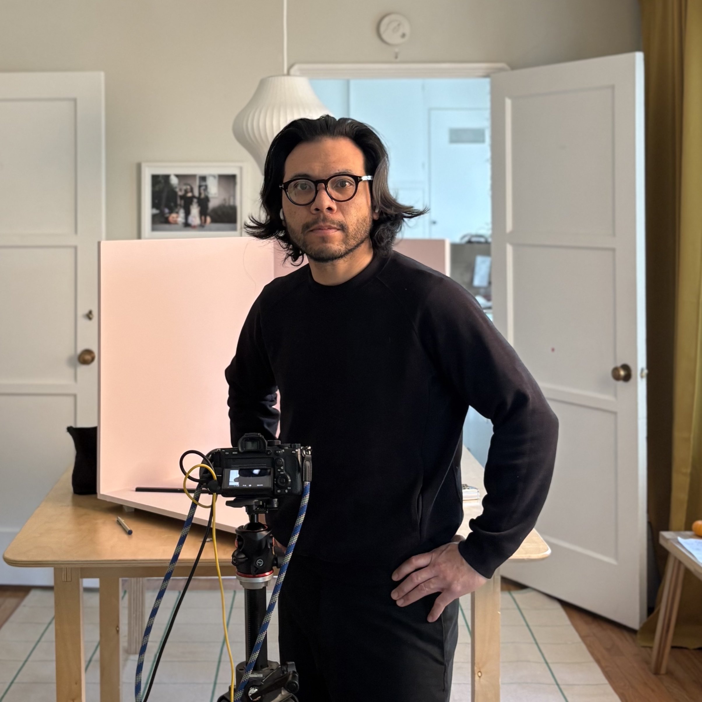 A man with glasses and long dark hair, wearing a black shirt, stands in a room with open doors. He is next to a camera on a tripod, which is directed towards a white, curved panel on a wooden table. The room has white walls, a photo in a frame, and part of a lamp visible.