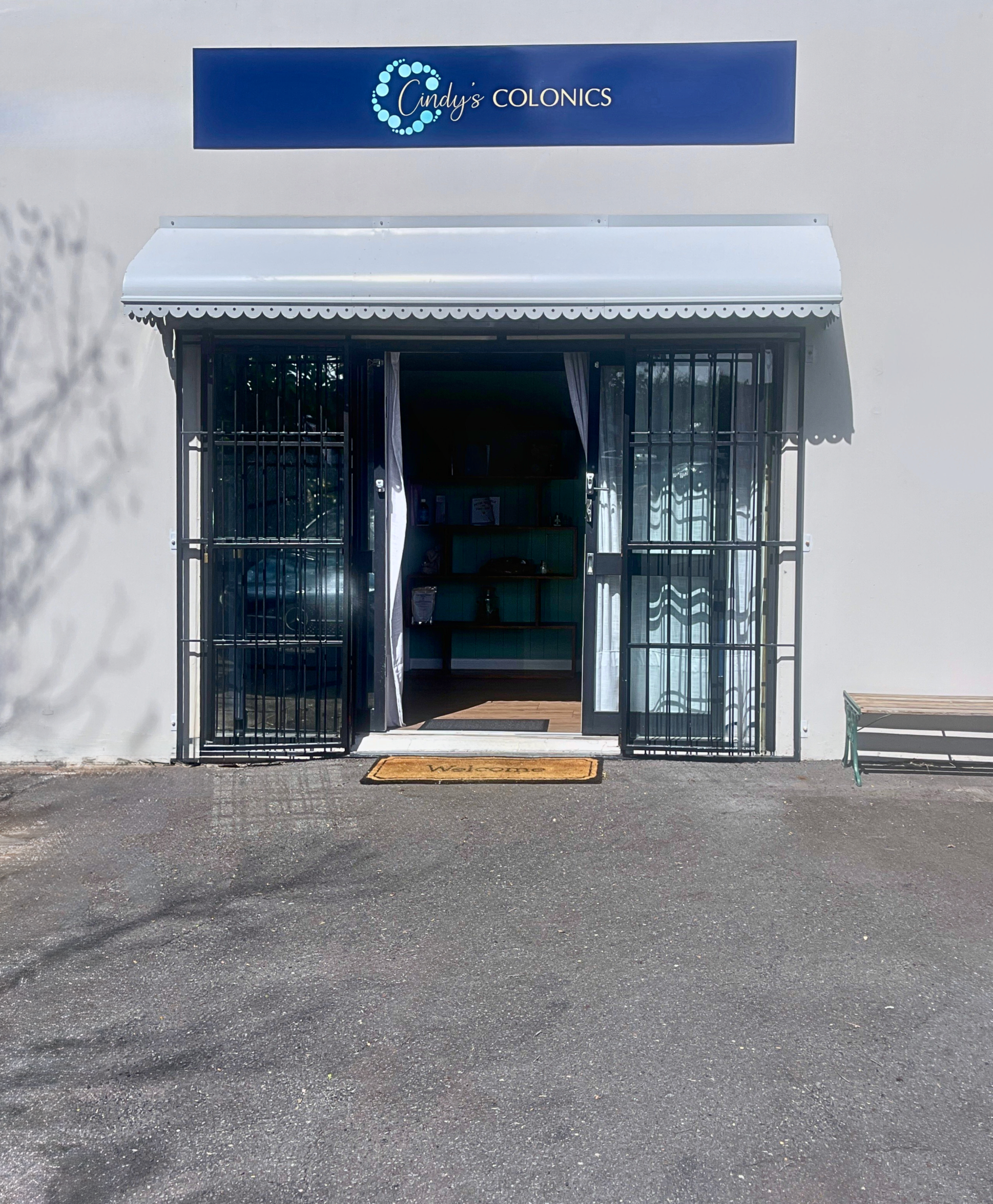 Front entrance of Cindy's Colonics with open gate, welcome mat, and white exterior wall, bench on the right, and sign above the doorway.