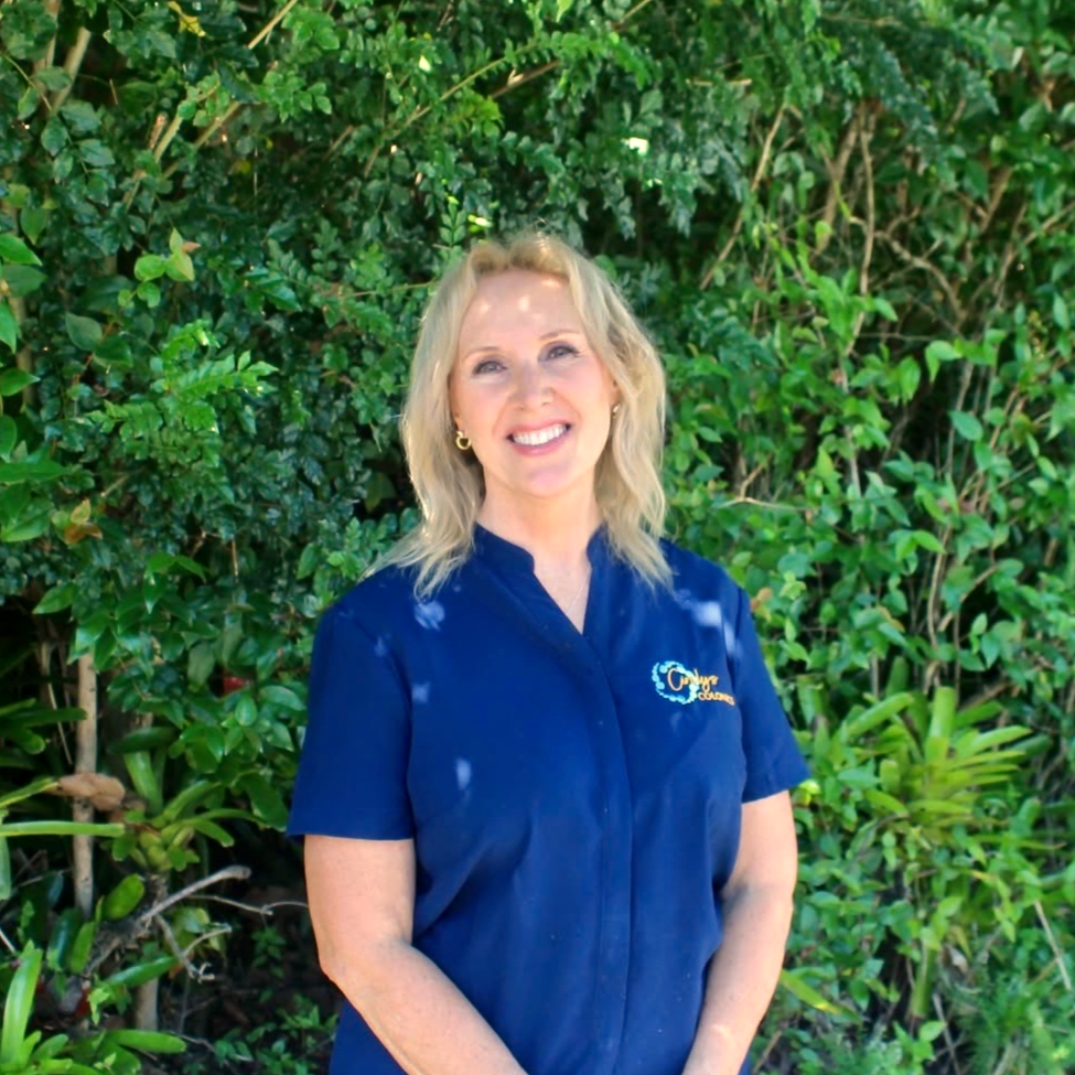 A woman with blonde hair, wearing a navy blue shirt with a logo, standing outdoors in front of green bushes, smiling at the camera.