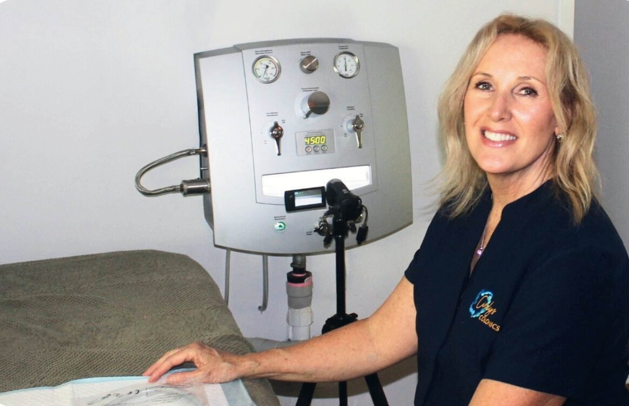 A woman in a black shirt with a logo on it sitting next to a medical device used for colonic hydrotherapy, smiling at the camera.